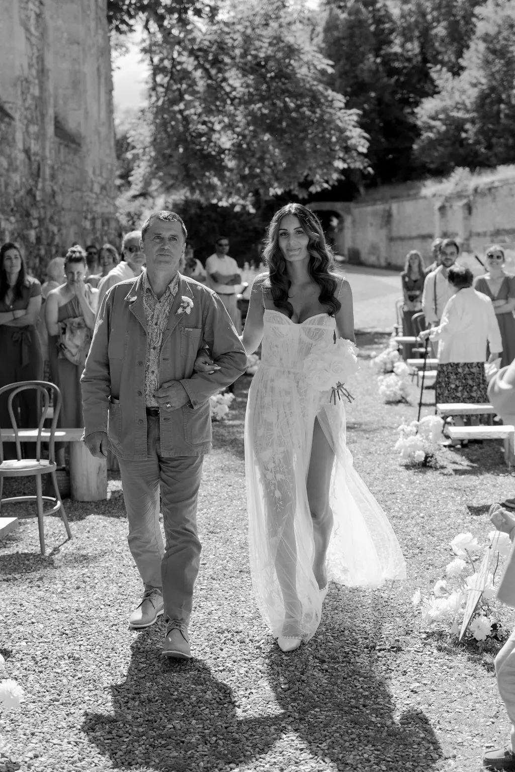Black-and-white processional of bride walking down gravel aisle in outdoor courtyard with seated guests