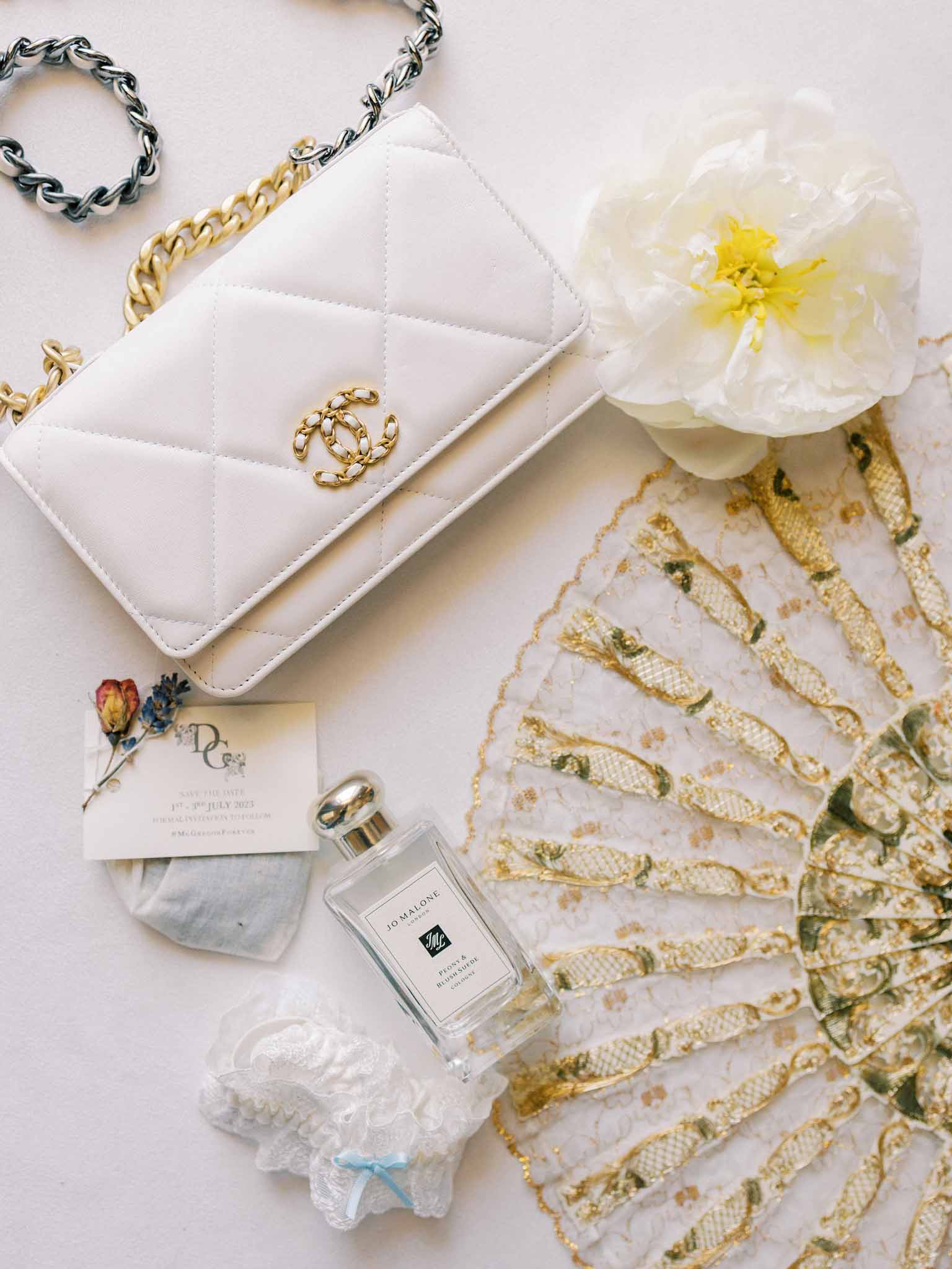 Flat lay of bridal accessories: white quilted handbag, lace garter, Jo Malone perfume, peony and gold chain bracelet
