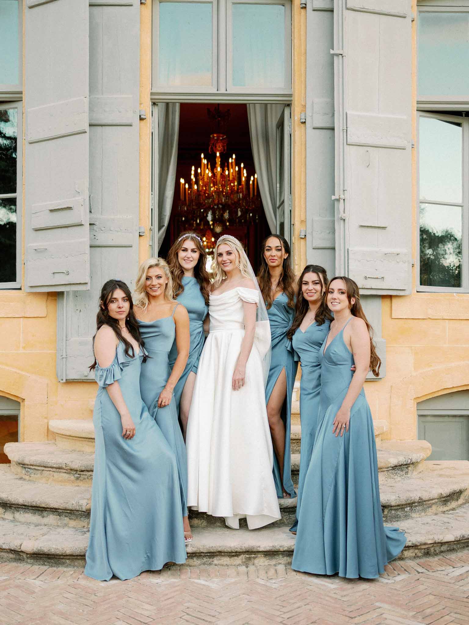 Bride with six bridesmaids in dusty blue gowns at chateau entrance with lit candelabra