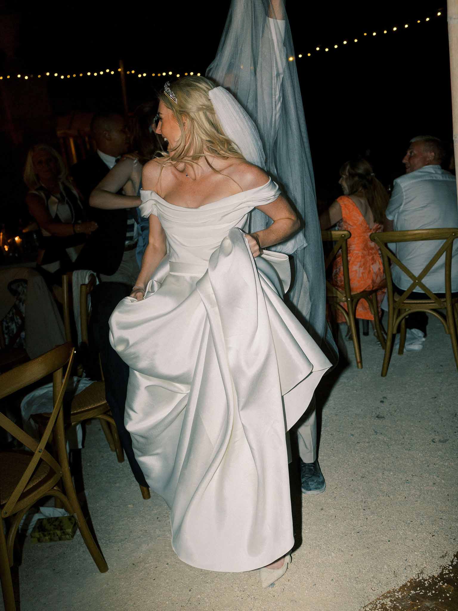 Bride in off-shoulder ivory gown with dusty blue veil dancing at outdoor reception under string lights at night