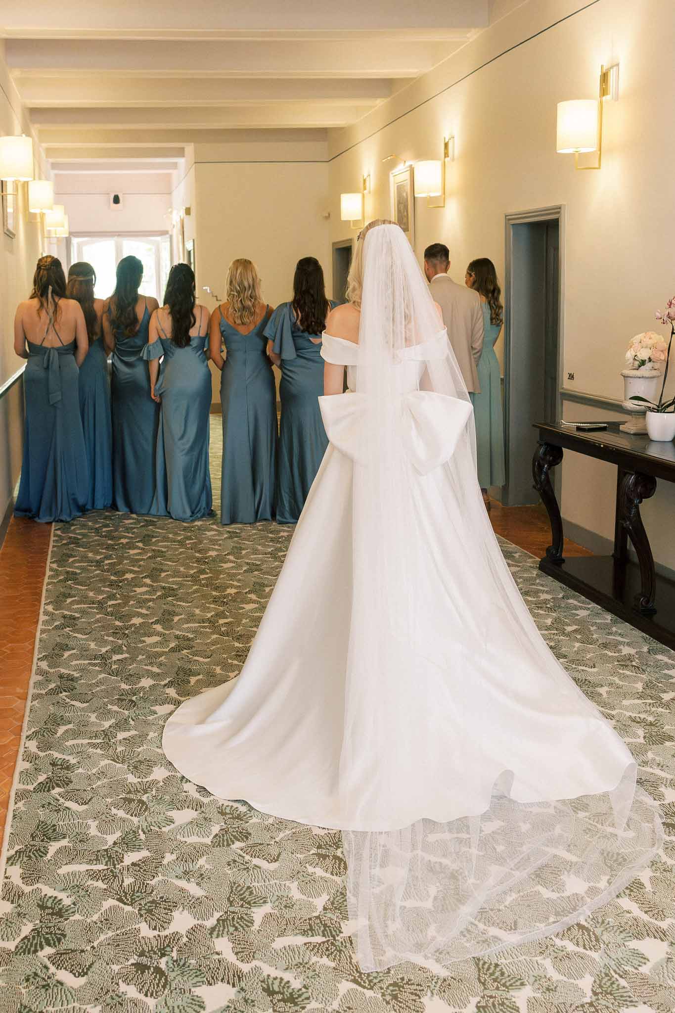 Bride in ivory gown seen from behind in elegant hallway as bridesmaids in slate blue dresses walk ahead