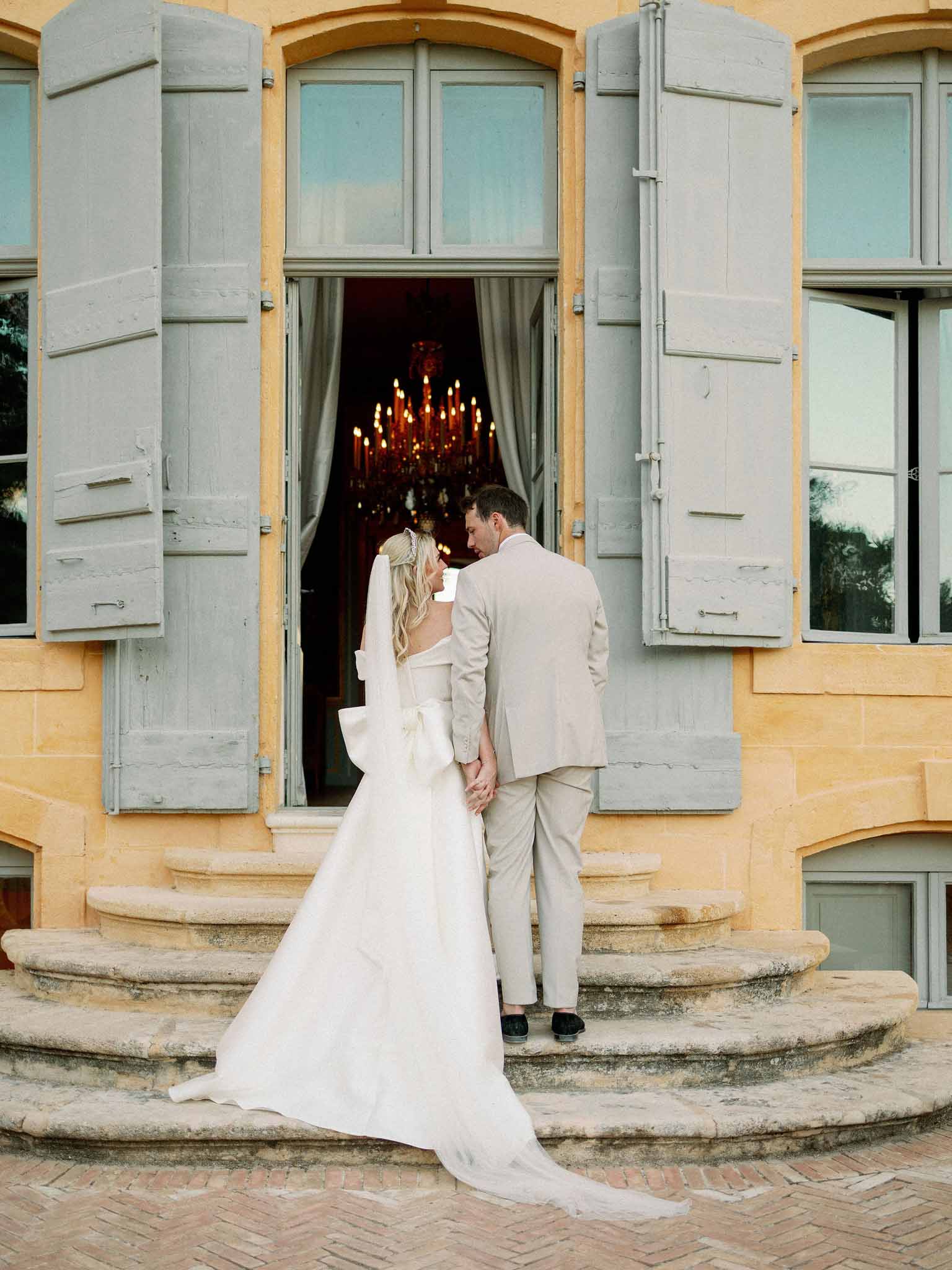 Bride and groom standing on curved stone staircase at château entrance with bronze chandelier visible inside