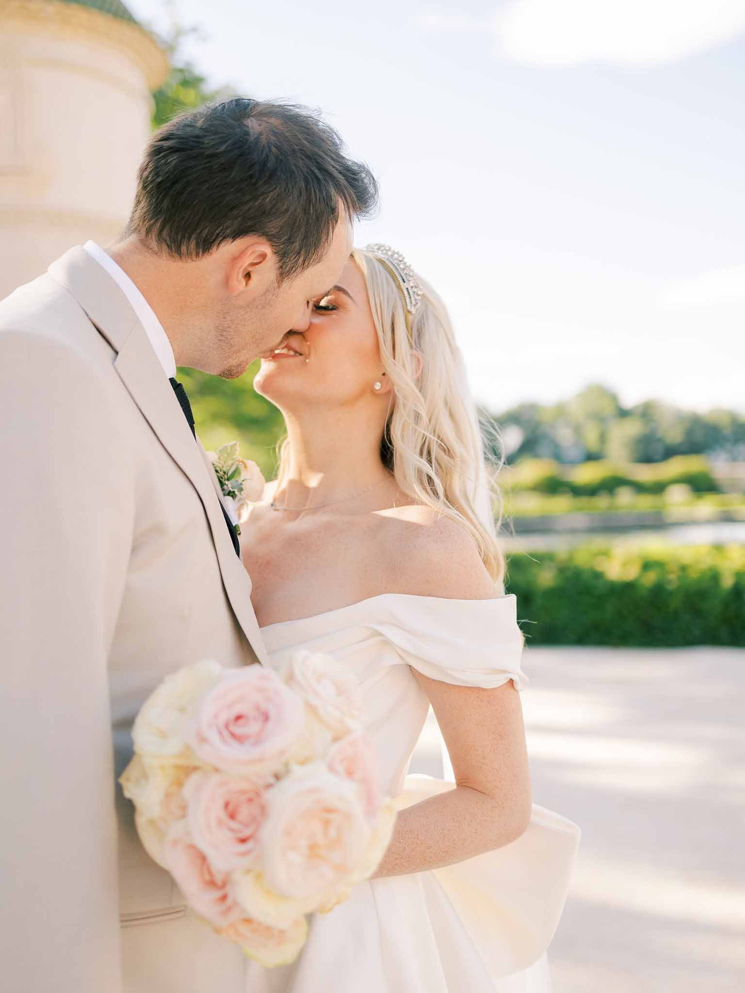 Groom in beige suit kissing bride on cheek as she holds blush and cream rose bouquet by riverside