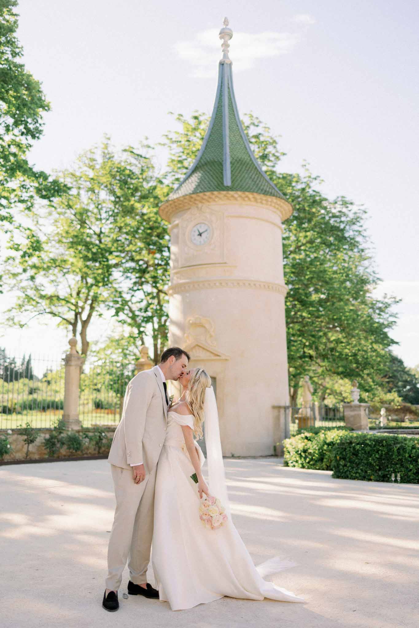 Bride and groom kissing in front of stone tower with green roof and manicured garden courtyard