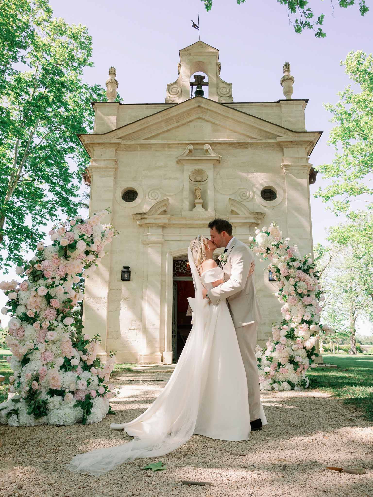 Groom lifts bride for a kiss outside stone chapel flanked by blush and cream rose floral installations