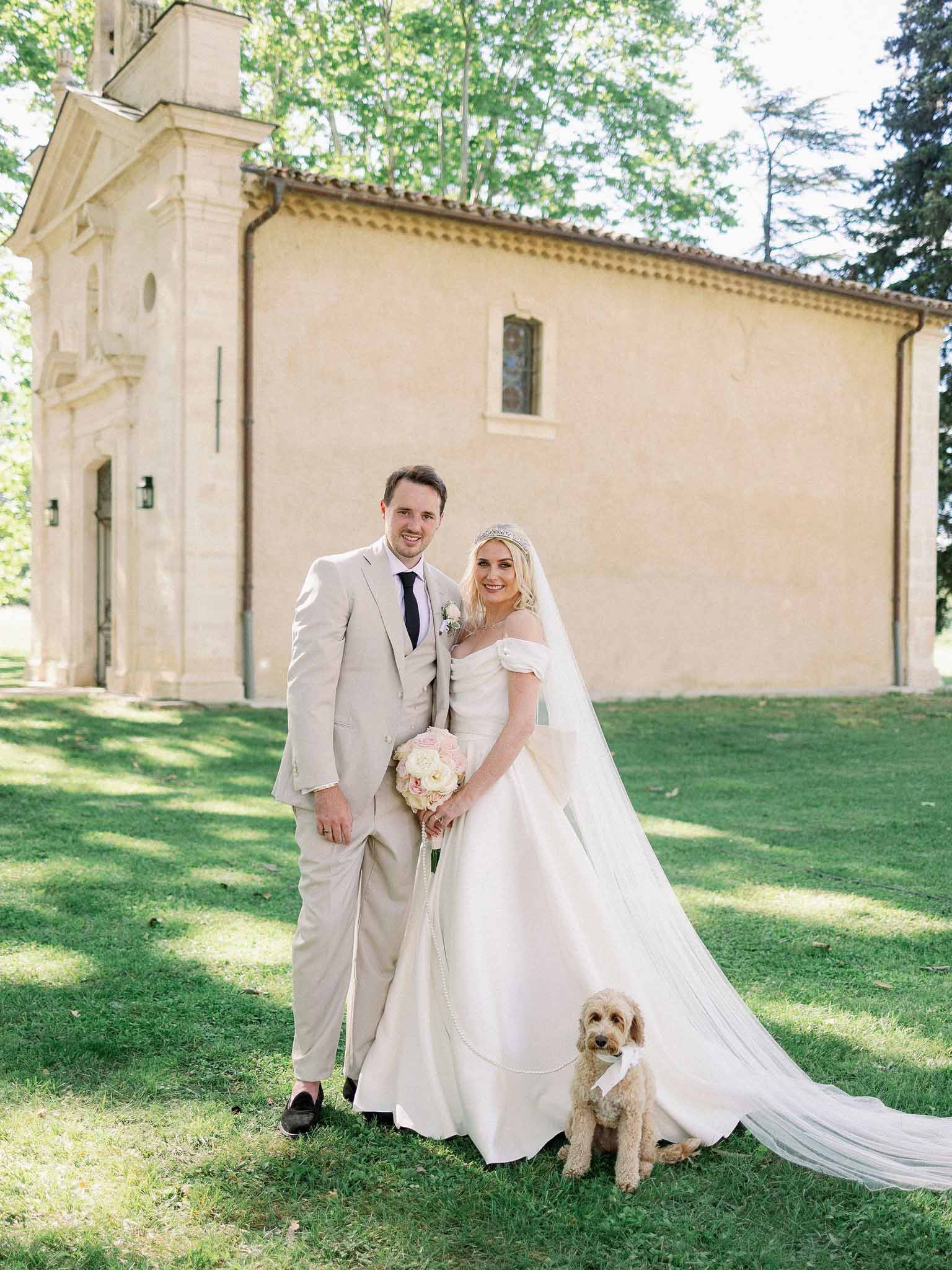 Bride and groom with small dog posing on lawn in front of stone chapel with ivy-covered roofline