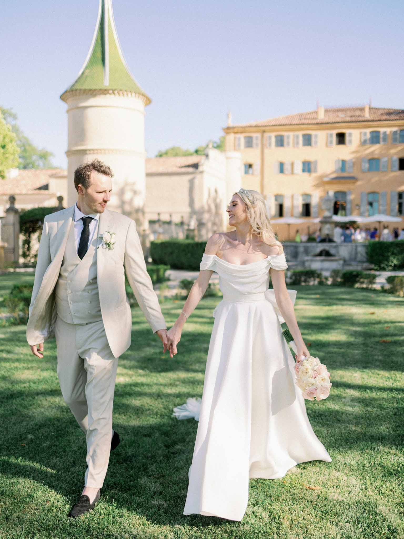 Bride and groom walk hand in hand across lawn with green-roofed château turret and formal gardens behind