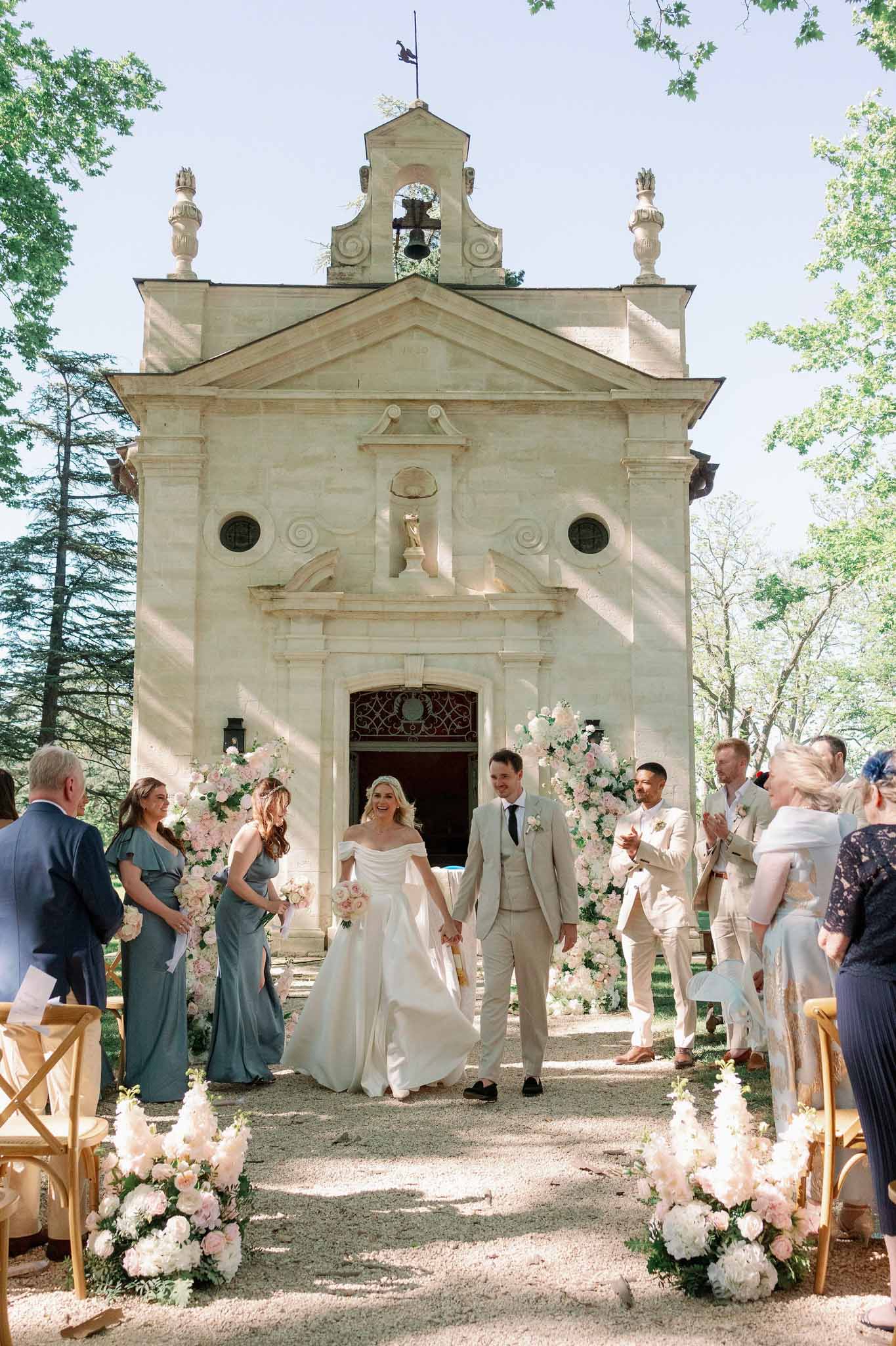 Bride and groom exiting stone chapel, bridesmaids in slate blue and groomsmen in cream suits flanking the aisle