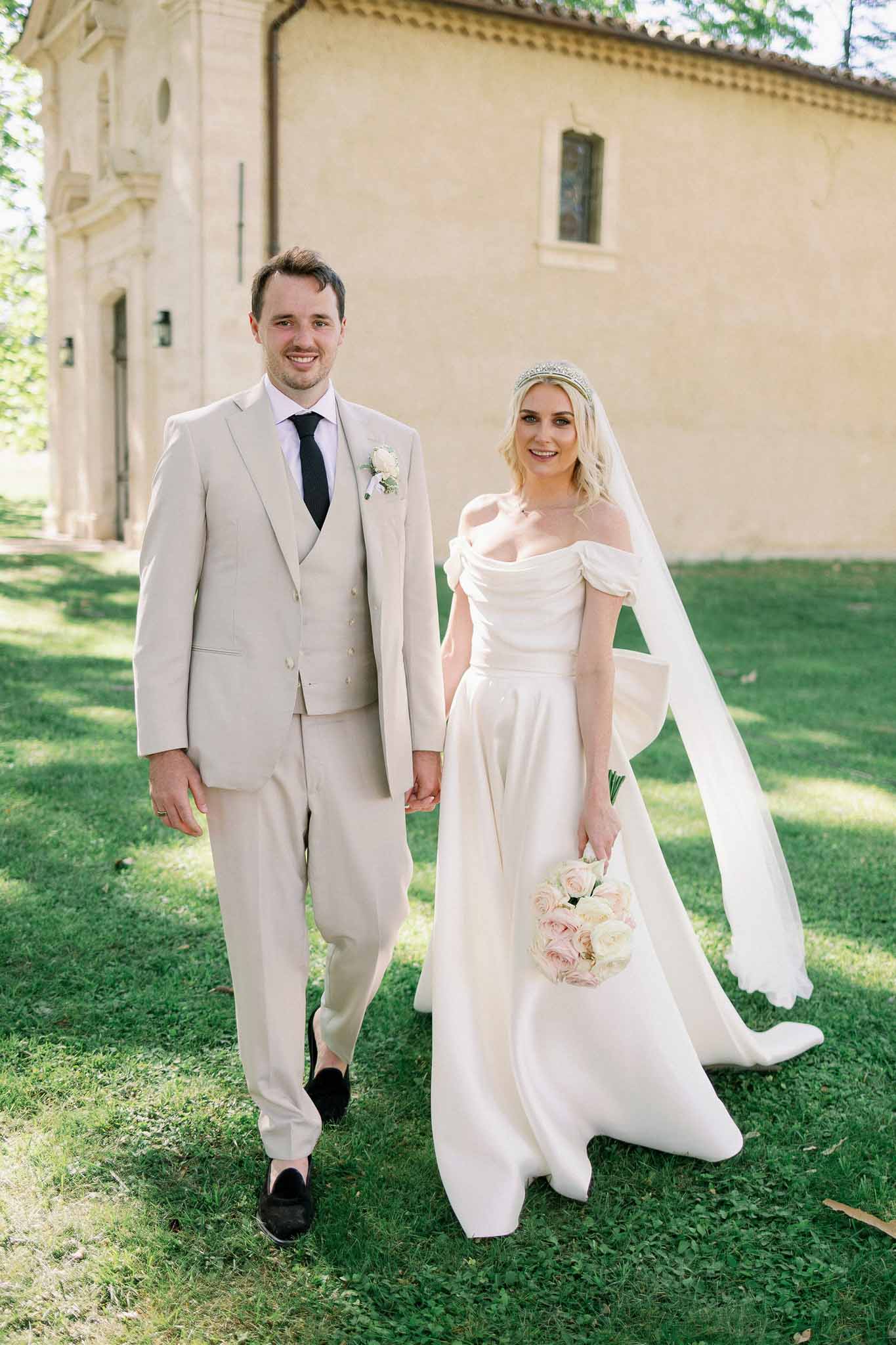 Bride and groom walking in front of pale yellow classical stone building, bride in ivory gown with blush bouquet