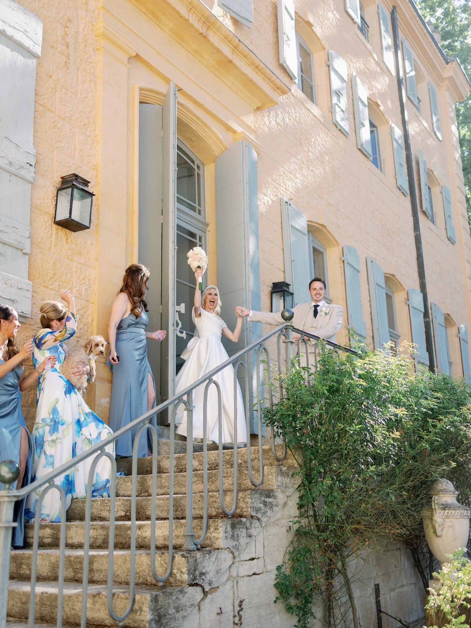 Bride and groom on stone staircase, bridesmaids in slate blue dresses and guests cheering below in bright daylight
