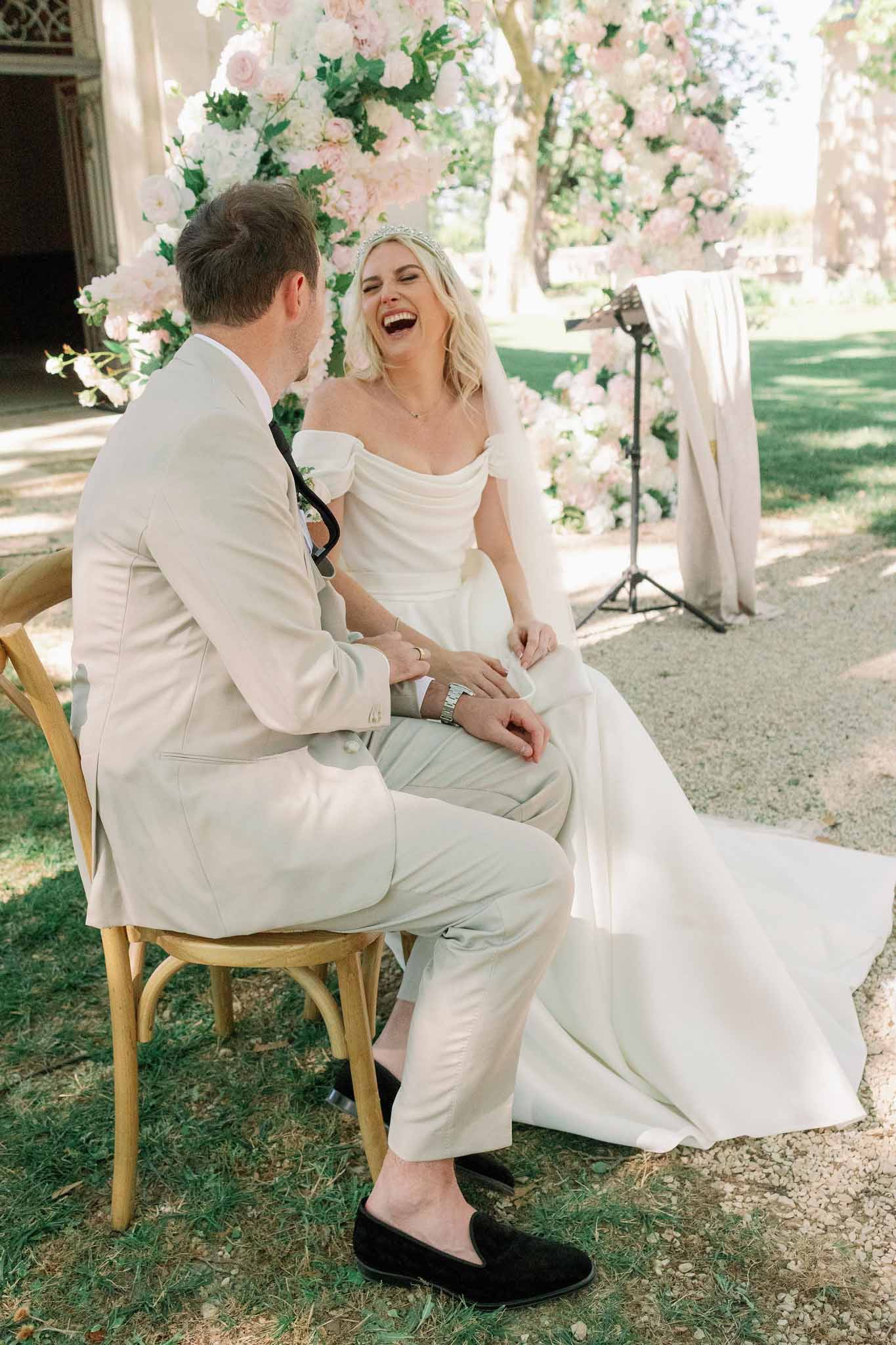 Bride and groom seated together laughing during outdoor ceremony with blush and white floral backdrop