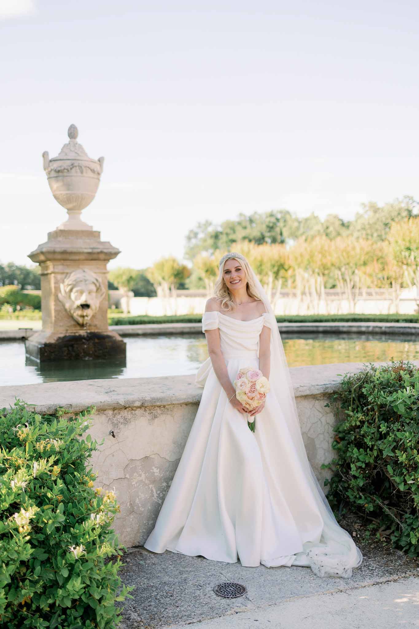 Bride in off-shoulder gown with blush and cream rose bouquet posing in formal garden beside stone canal