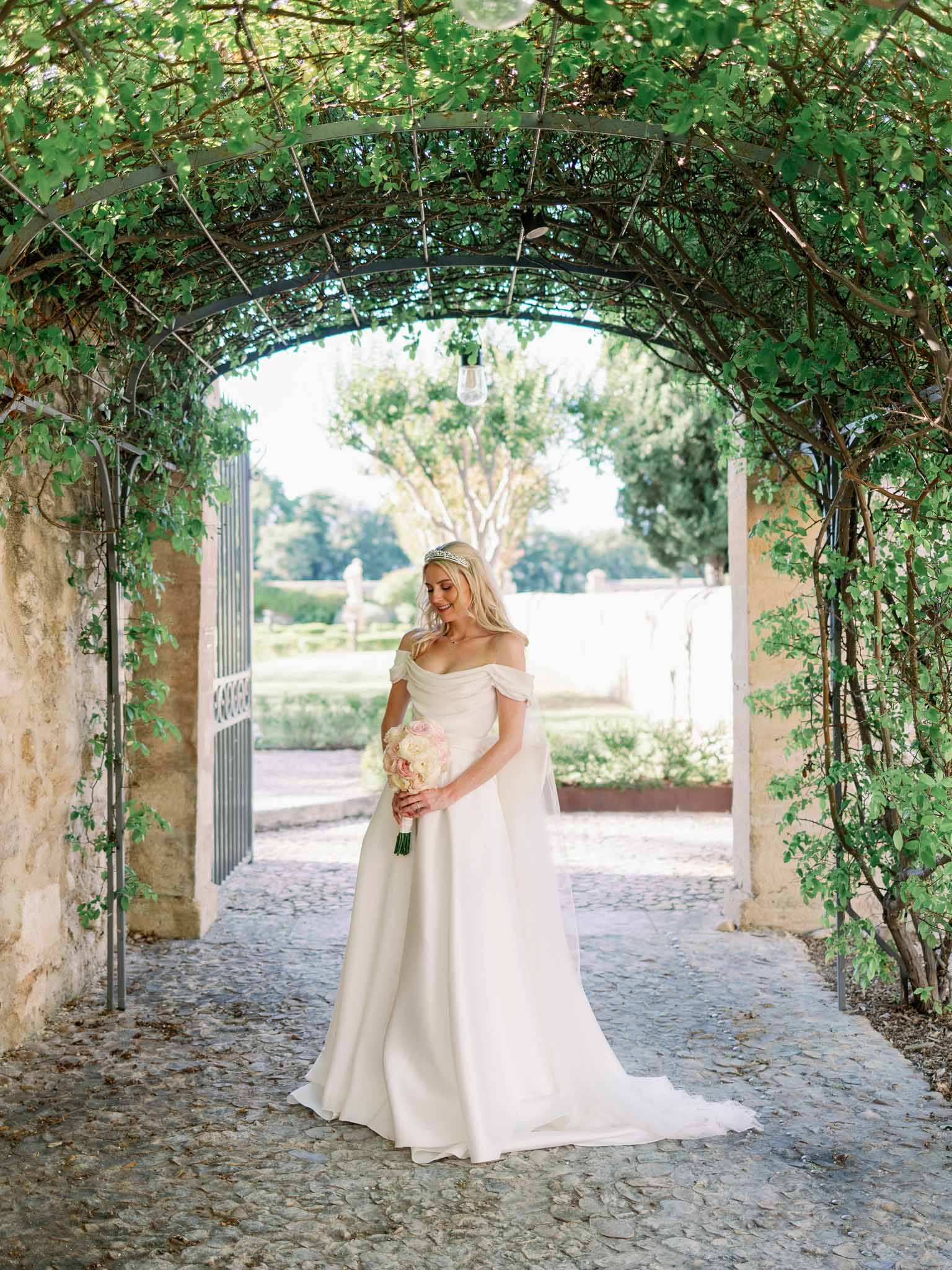 Bride in off-shoulder ivory silk gown with gold leaf crown holding bouquet beneath ivy-covered green metal arbor in stone garden passage