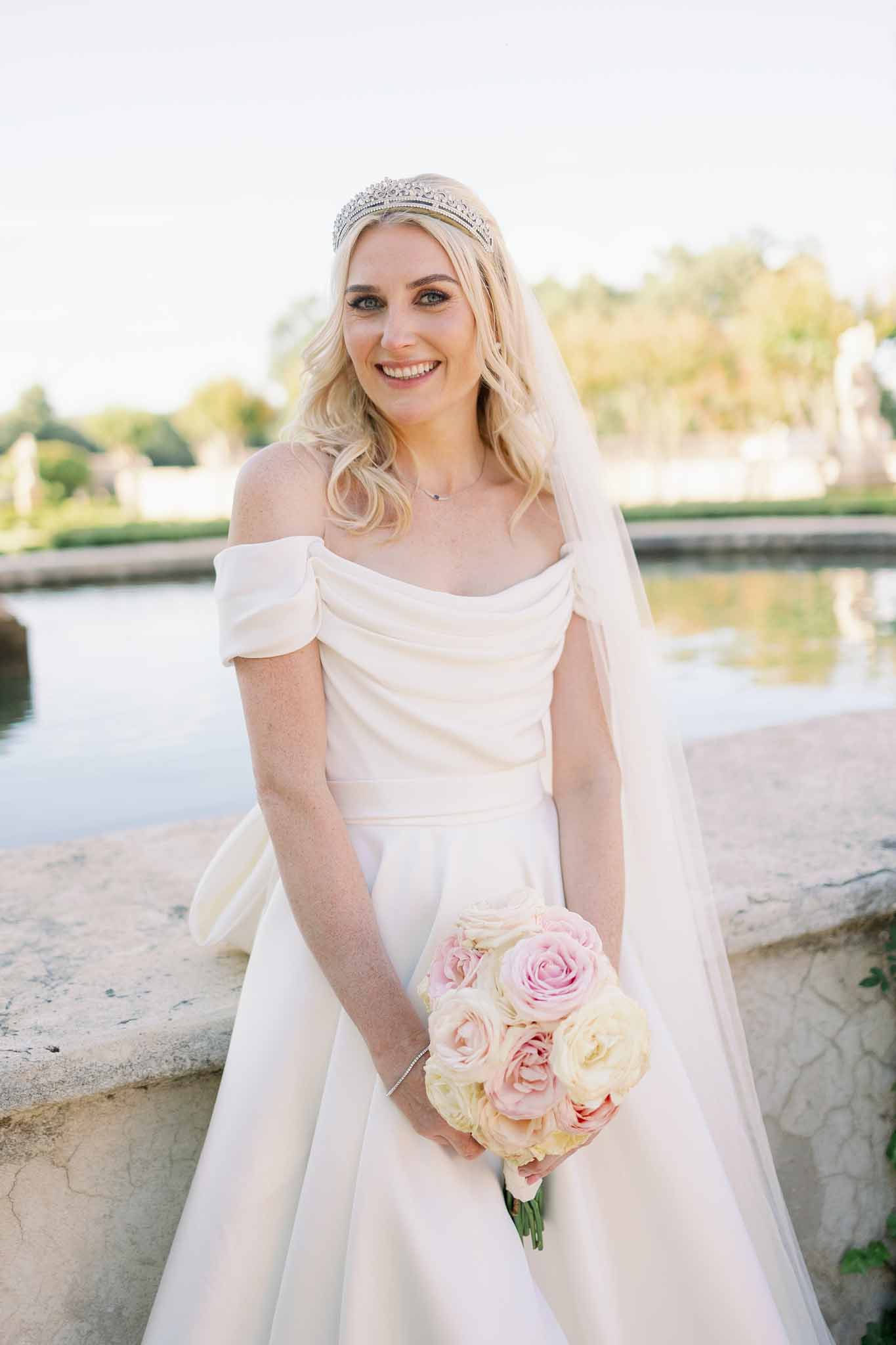 Bride in ivory off-shoulder dress with veil and beaded headpiece holding pink and cream rose bouquet by a lake