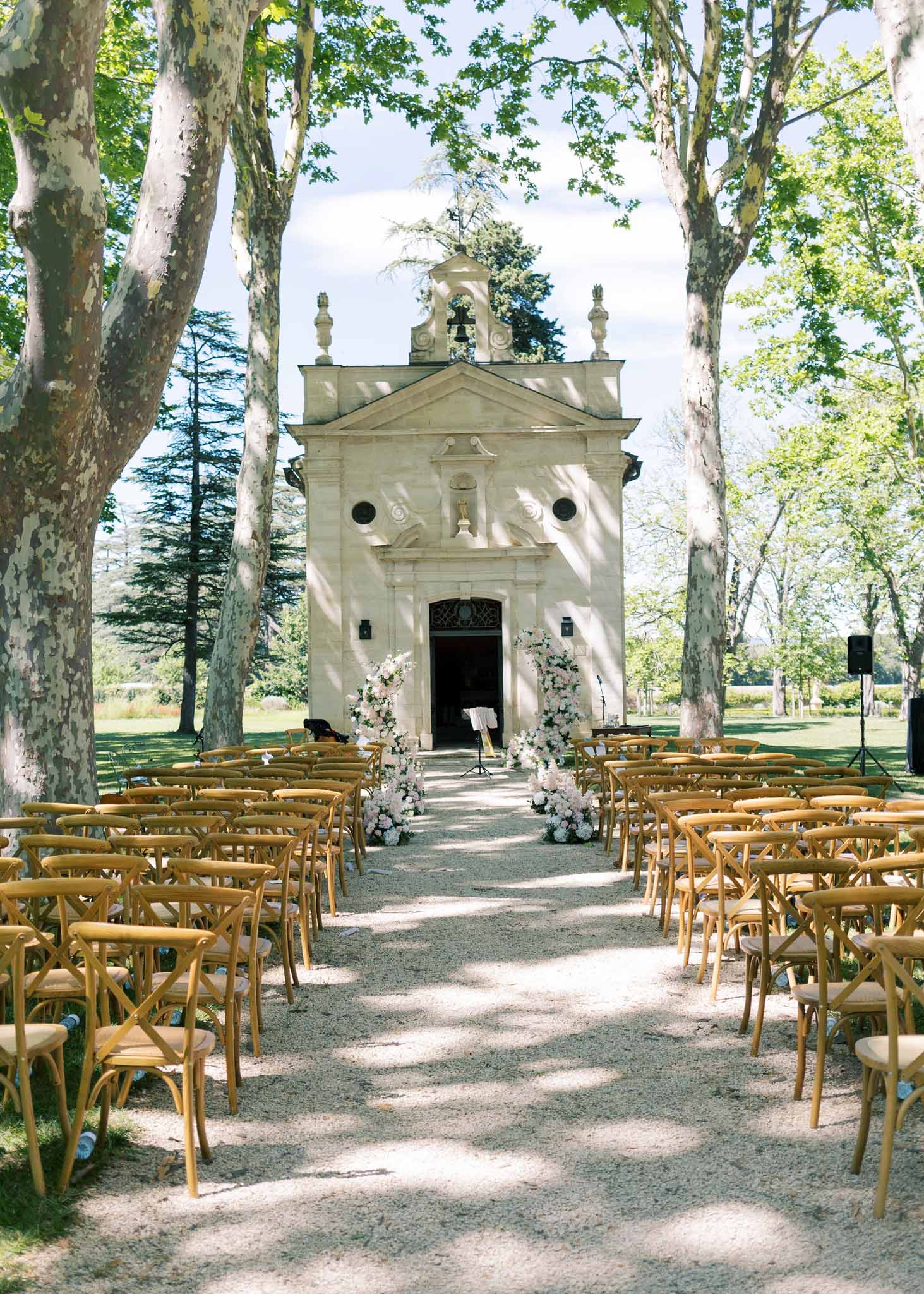 Outdoor ceremony setup in a chapel courtyard with wooden cross-back chairs and white floral urns at entrance