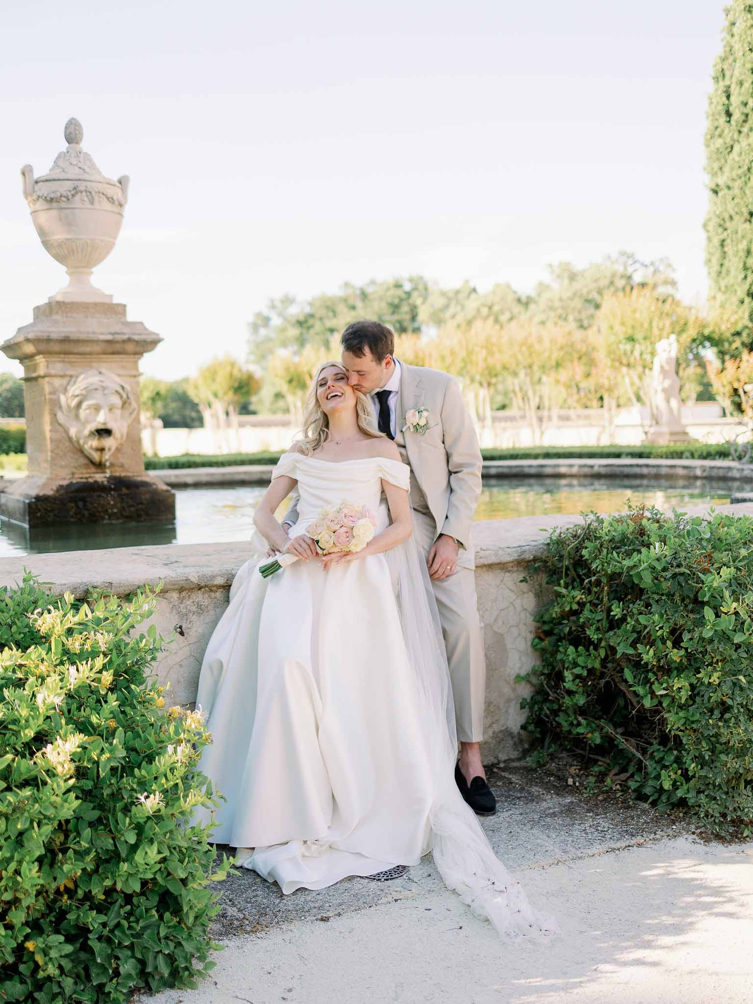 Bride in off-shoulder tulle gown and groom in taupe suit smile at each other beside formal garden reflecting pool