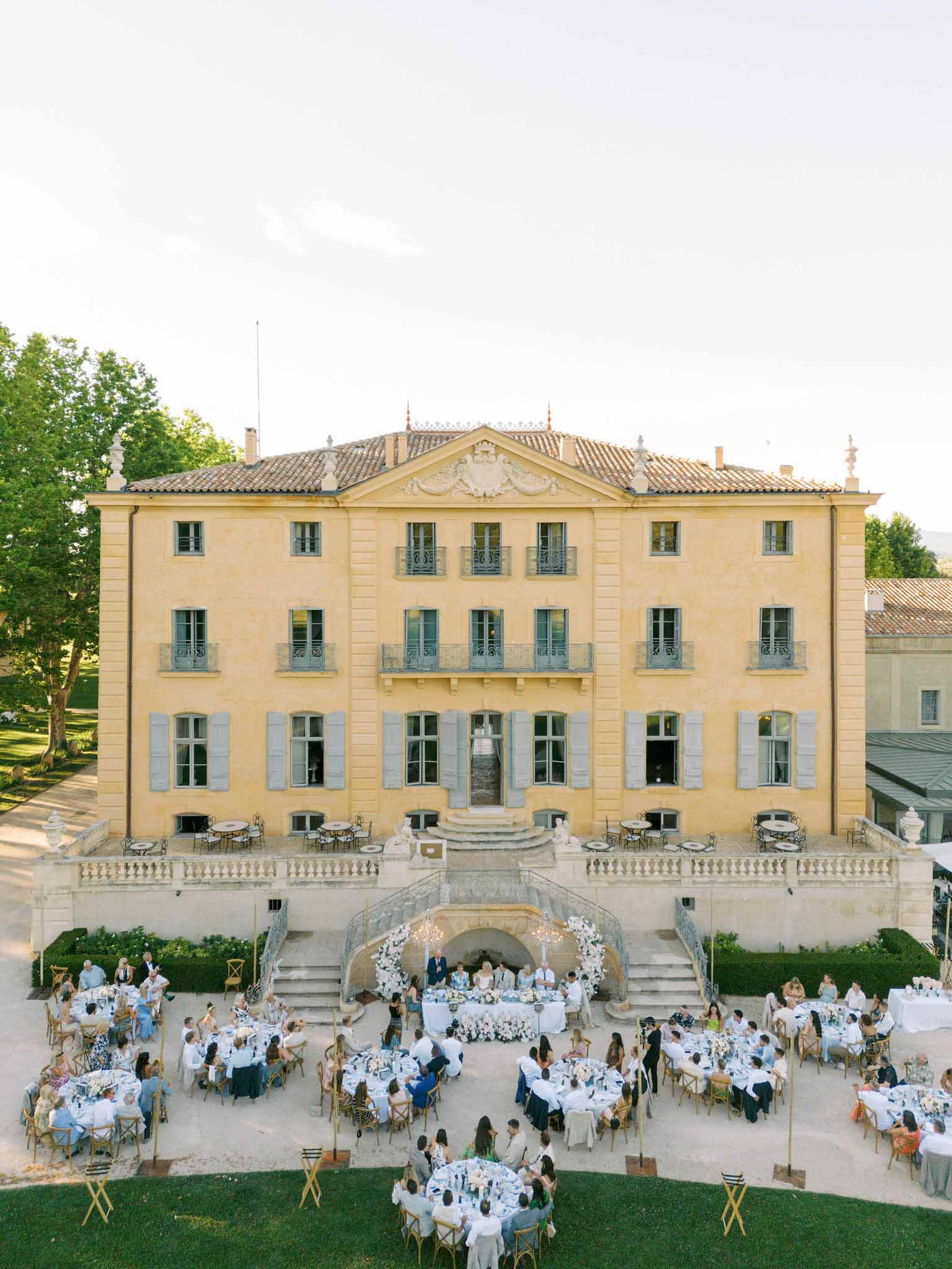 Aerial view of a neoclassical estate with 100+ guests at round white-linened tables across a terrace and lawn during a daytime reception.