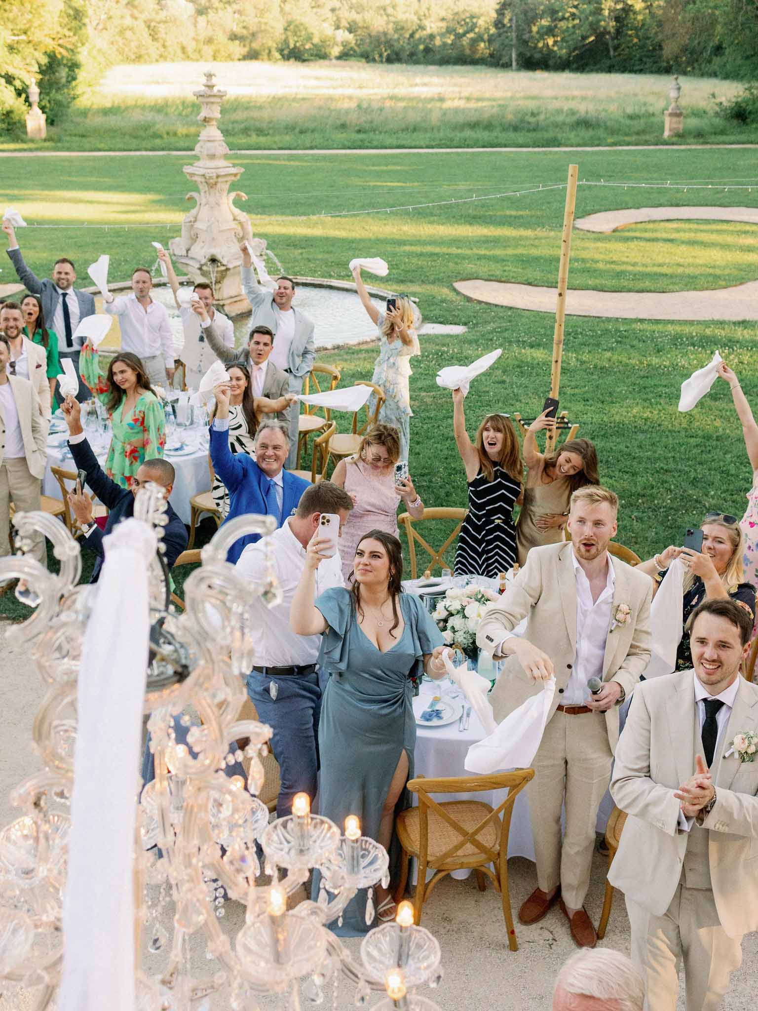 Wedding guests toss white papers in the air during outdoor reception on estate lawn with crystal chandeliers and stone fountain