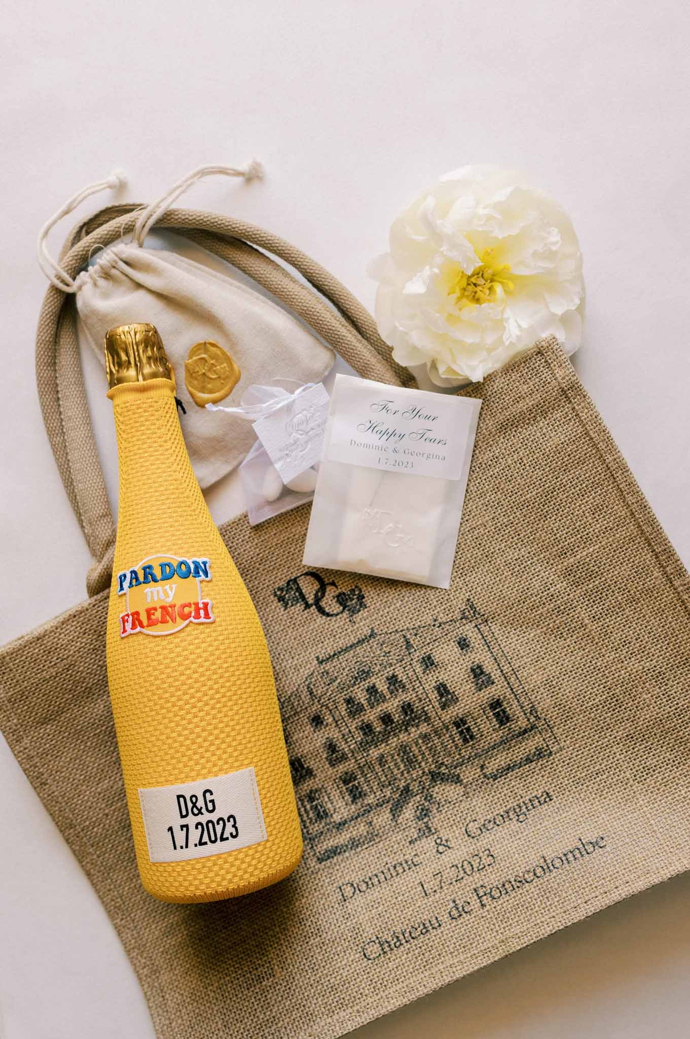 Flat lay of wedding favours on linen napkin: personalized champagne bottle, gold wax seal, cream pouch and a white peony.