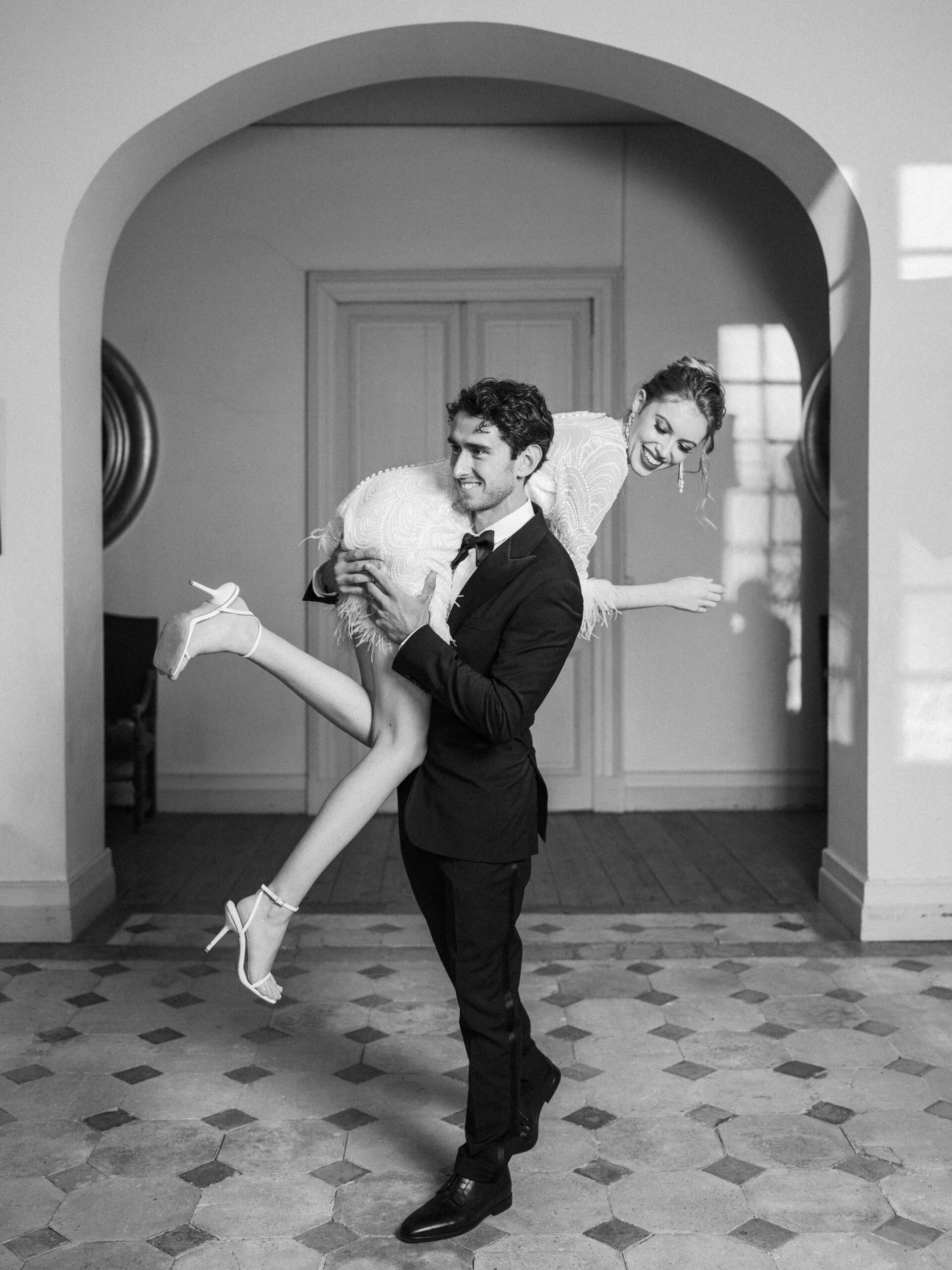 Black-and-white full-length portrait of a couple inside a chateau or manor house, with the groom carrying the bride over his shoulder in a playful pose. The groom wears a dark fitted suit with a bow tie and dress shoes, while the bride wears a short heavily embellished mini dress featuring feather trim at the hem, paired with white strappy heeled sandals and long drop earrings. The interior setting features a large arched doorway, panelled doors, parquet wood flooring visible through the arch, and a geometric diamond-and-hexagon patterned tile floor in the foreground. The image is shot in high contrast black and white with bright light entering from windows in the background, and the styling has a modern, fashion-forward aesthetic.