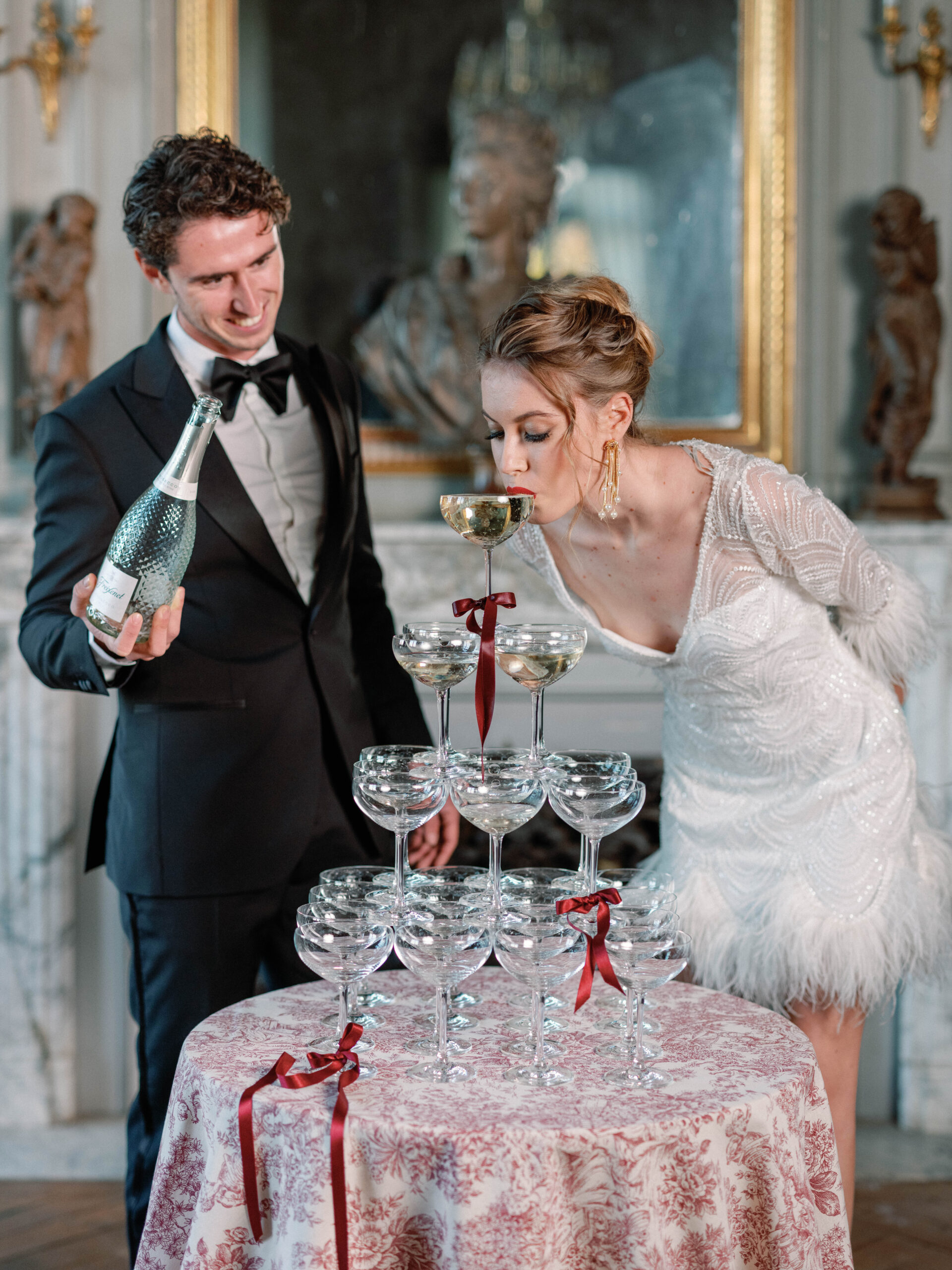 A couple interacts with a champagne tower during what appears to be a reception moment inside an ornate French château or palace interior. The groom wears a black tuxedo with a black bow tie and holds a champagne bottle, while the bride leans forward to sip directly from the top coupe glass of the tower. The bride wears a short white beaded and feather-trimmed dress with long beaded sleeves, statement gold drop earrings, and a red lip. The champagne tower is built from coupe glasses adorned with deep burgundy satin ribbon bows, displayed on a round table draped in a red and ivory toile de Jouy linen, also tied with a burgundy ribbon. The background features white ornamental paneling, gold-framed mirrors, gilded wall sconces, and decorative classical busts, pointing to a grand French interior. The overall styling is 1920s Art Deco-inspired with a rich burgundy and ivory color palette. Medium portrait-style shot.