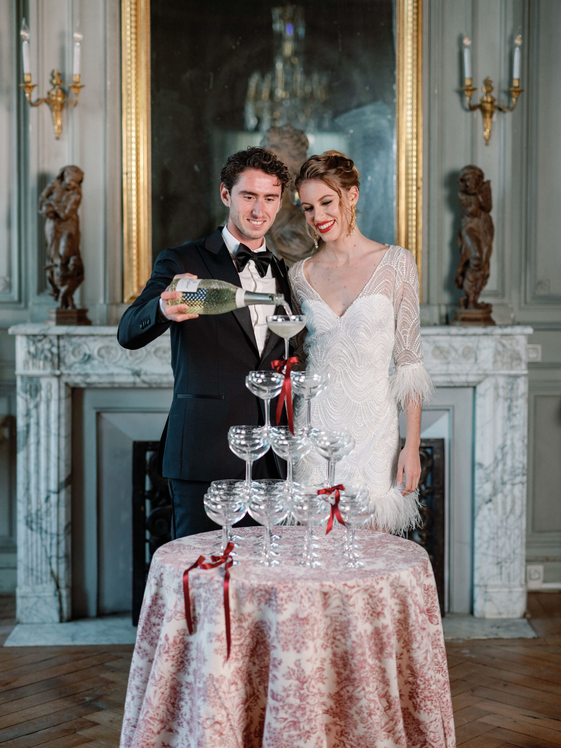 A couple performs a champagne tower pour during a wedding reception inside what appears to be a French chateau or palace-style venue. The groom wears a navy tuxedo with black bow tie, while the bride wears a fitted white beaded gown with feather-trimmed sleeves and gold drop earrings, her hair styled in an updo. The champagne coupe tower sits on a round table covered in a red-and-cream toile de Jouy linen, with small red satin ribbon bows tied to the glasses. The room features a white marble fireplace, gilt-framed antique mirror, bronze wall sconces with candles, and decorative bronze figural sculptures, all set against grey-painted paneled walls with parquet flooring. The overall styling theme is classic French with Art Deco-influenced bridal fashion. Medium portrait shot framing both subjects from the waist up.