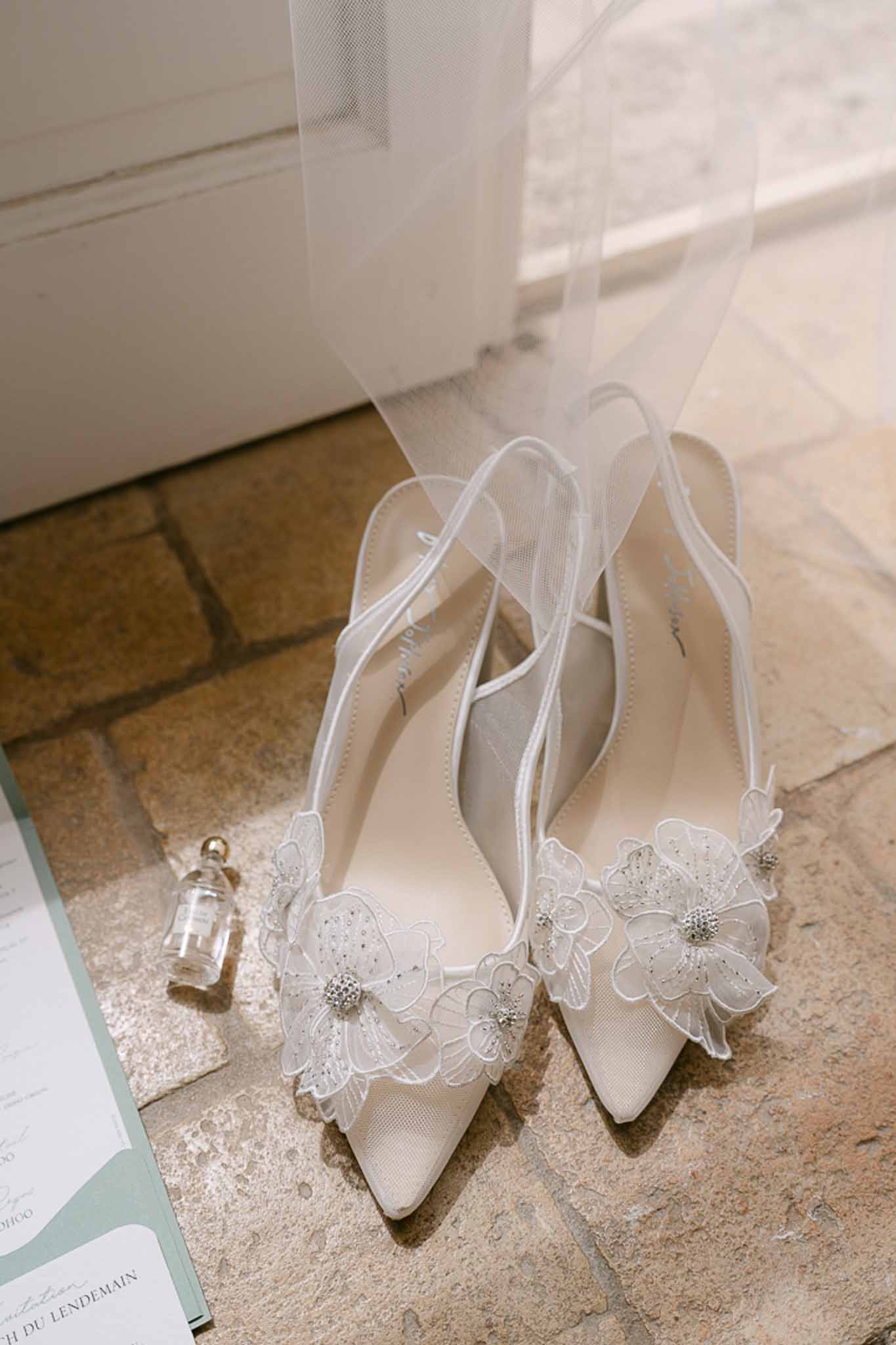 Close-up of bridal ivory heels with white lace floral appliqués, crystal beading and tulle veil on stone floor