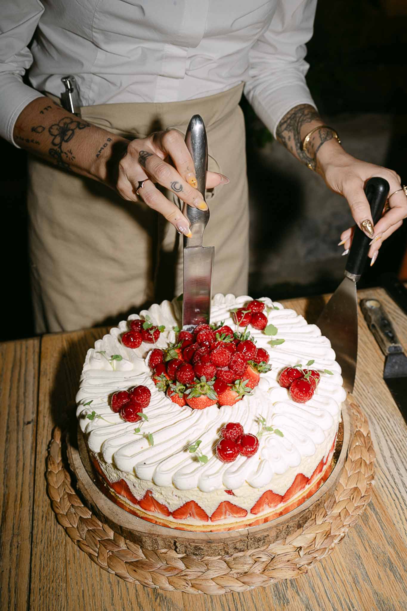 Bride and groom cut two-tier wedding cake topped with fresh strawberries and raspberries on wooden board