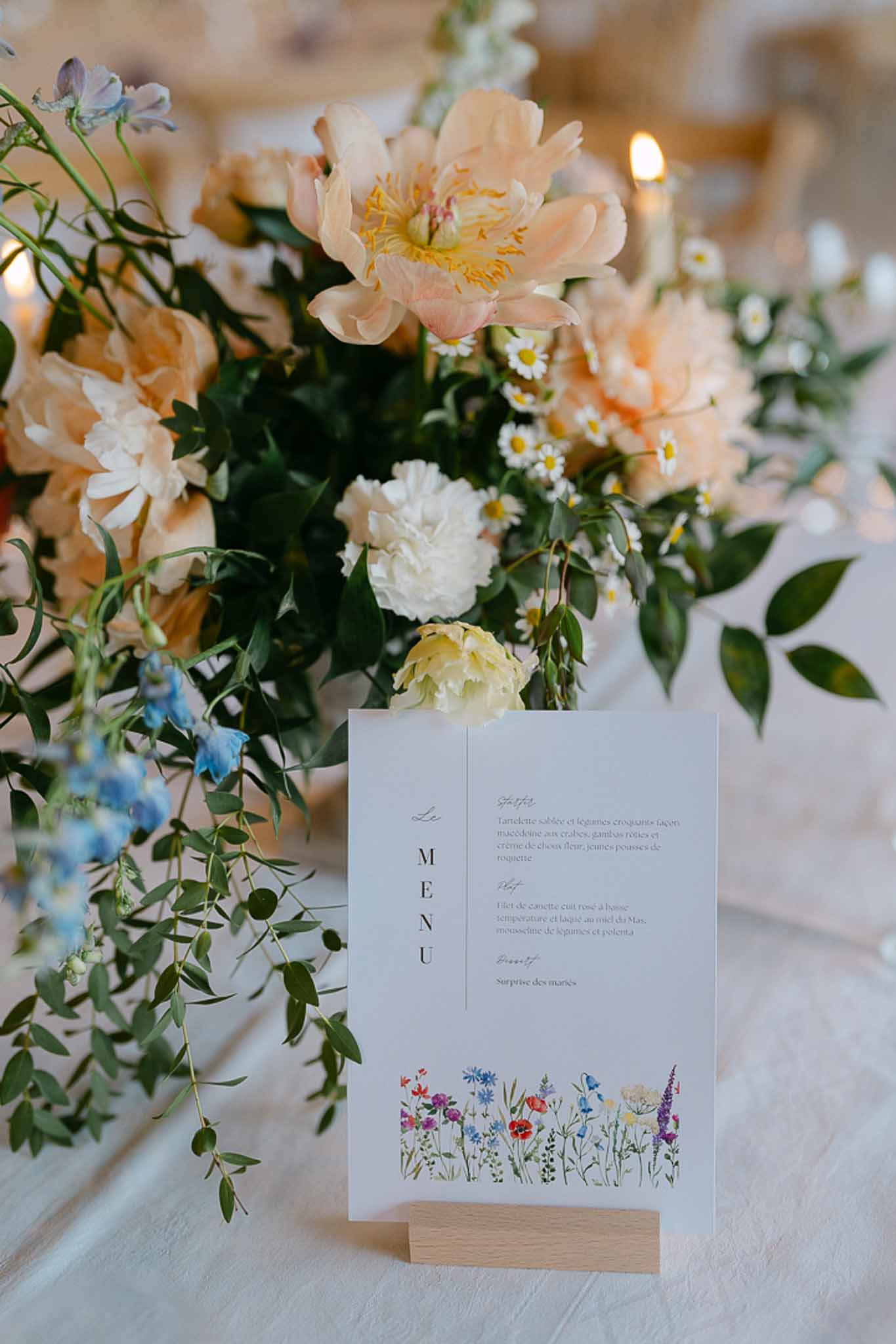 Reception table detail with menu card in wood stand alongside cream and blush peonies, delphiniums, and candlelit background