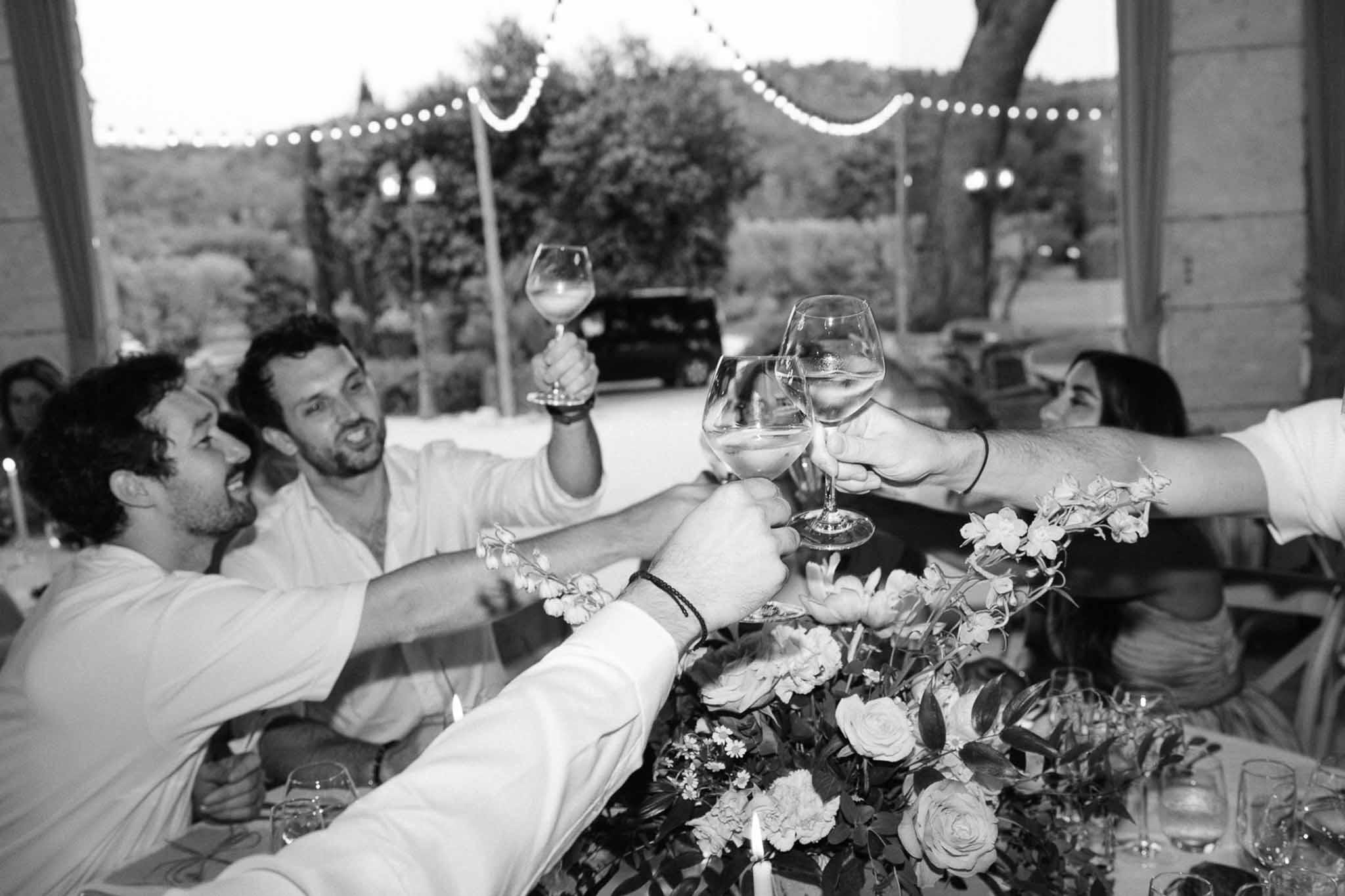 Black and white; multiple guests raising wine glasses in a toast at an outdoor table, string lights above and rose centerpiece.