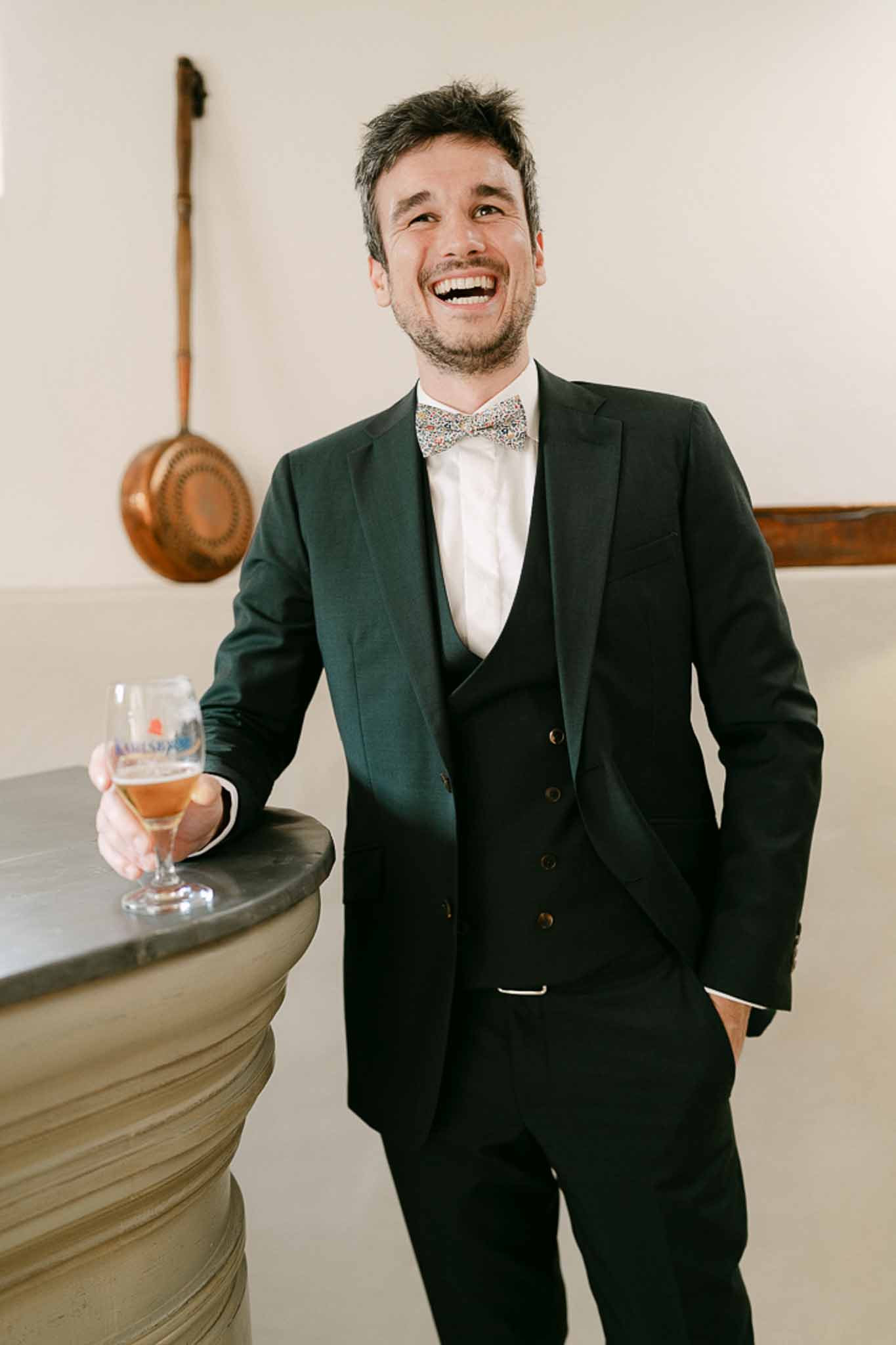 Groom in dark green double-breasted suit laughing indoors, holding a glass of amber beverage against a pale cream wall.