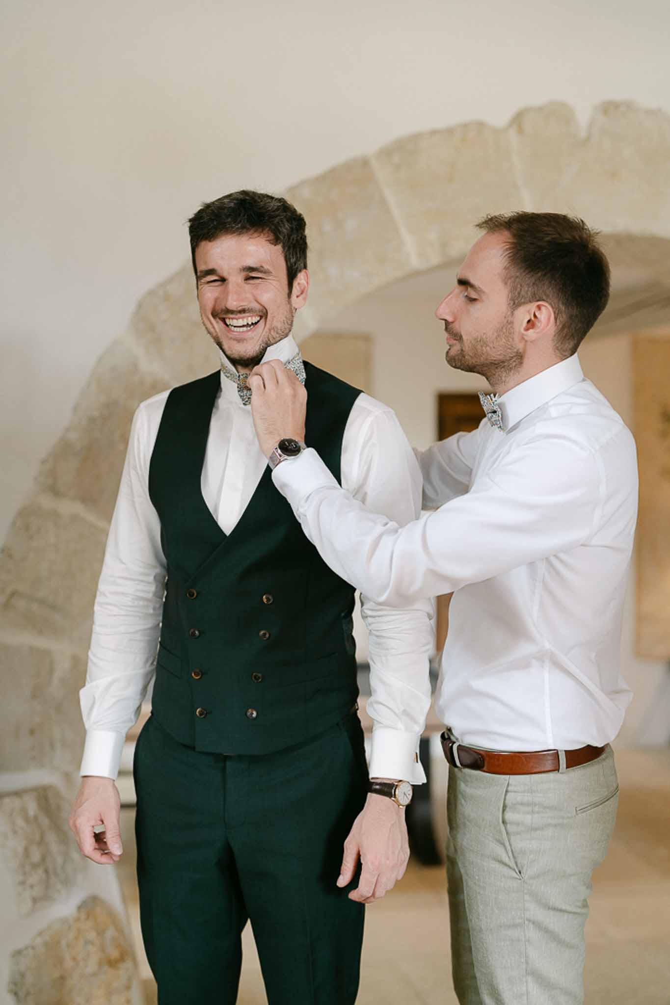 Groomsman adjusting groom's bow tie, groom in dark green double-breasted waistcoat in stone-walled room