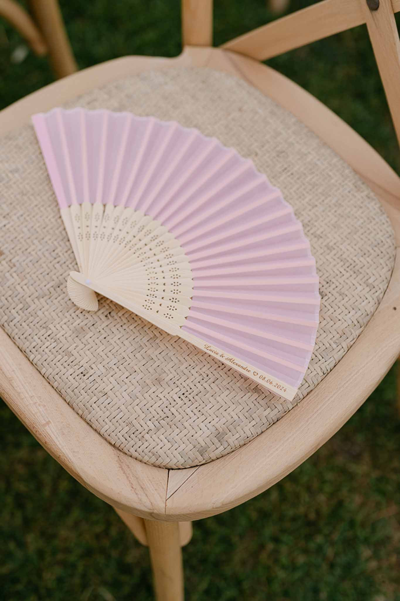 Pale lavender folding fan guest favour resting on wooden chair cushion at outdoor ceremony