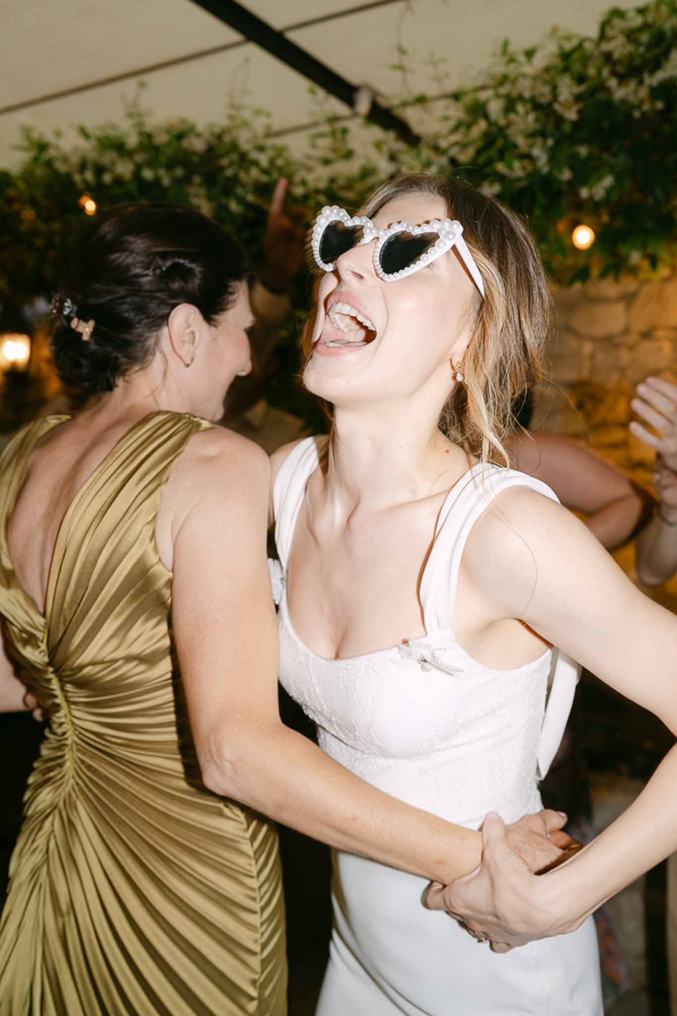 Two guests laughing under foliage garland and amber lights, one wearing heart-shaped novelty sunglasses at reception