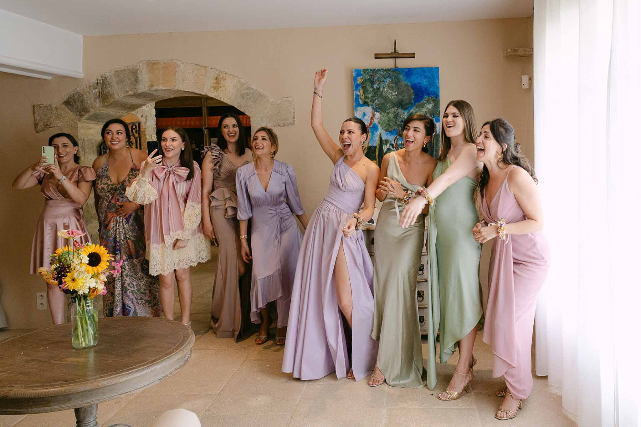 Bride in lavender wrap dress with nine bridesmaids in mixed pastel gowns celebrating in a stone-walled chapel space.