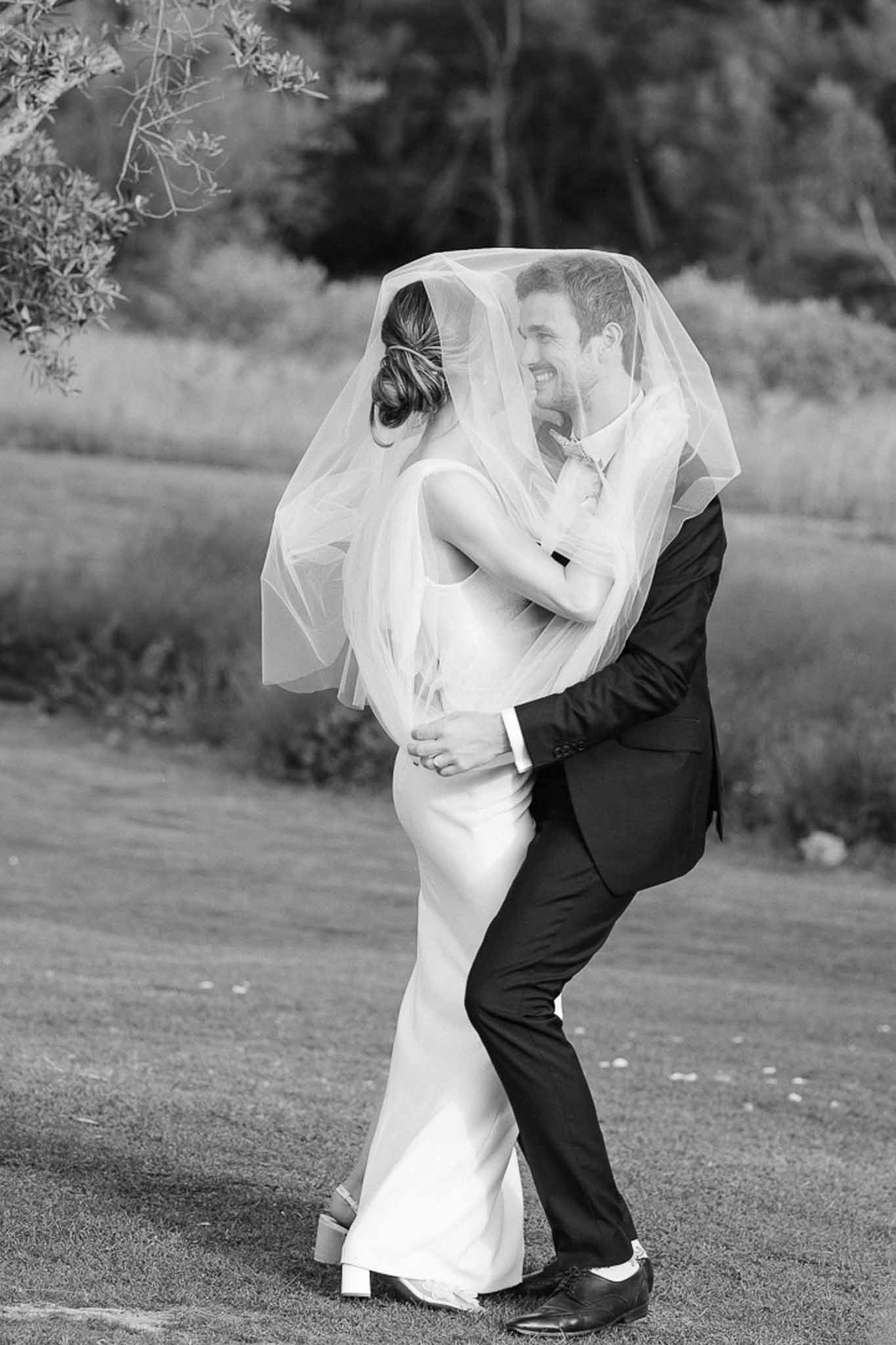 Black and white photo of groom lifting bride as her veil billows around them on a garden path