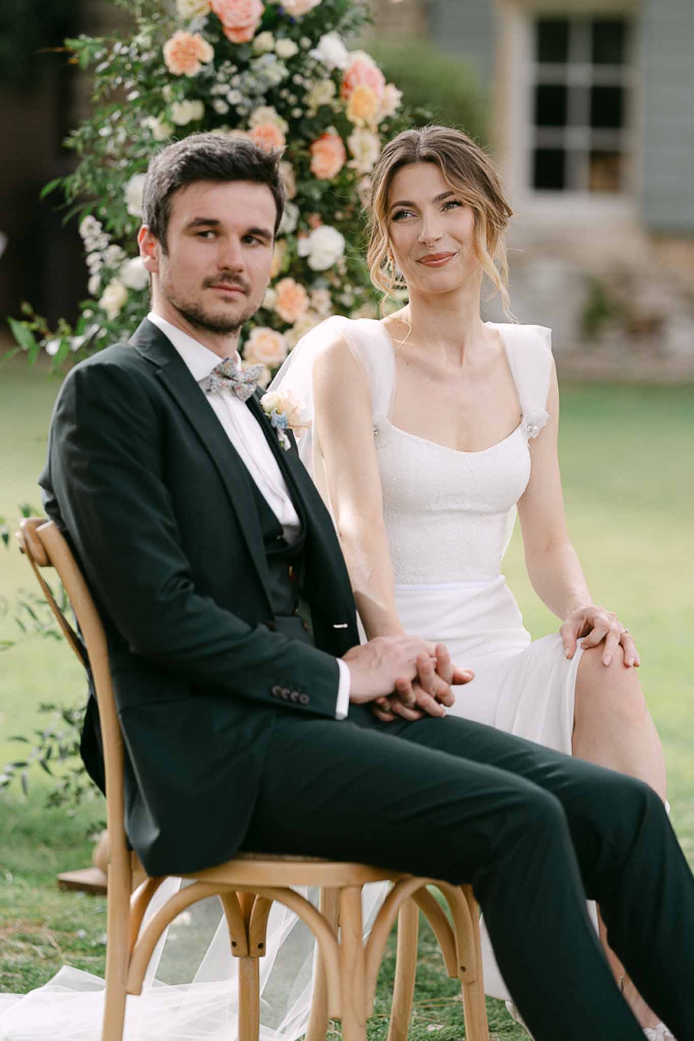 Bride and groom seated on bentwood chairs before coral and cream floral arch in garden