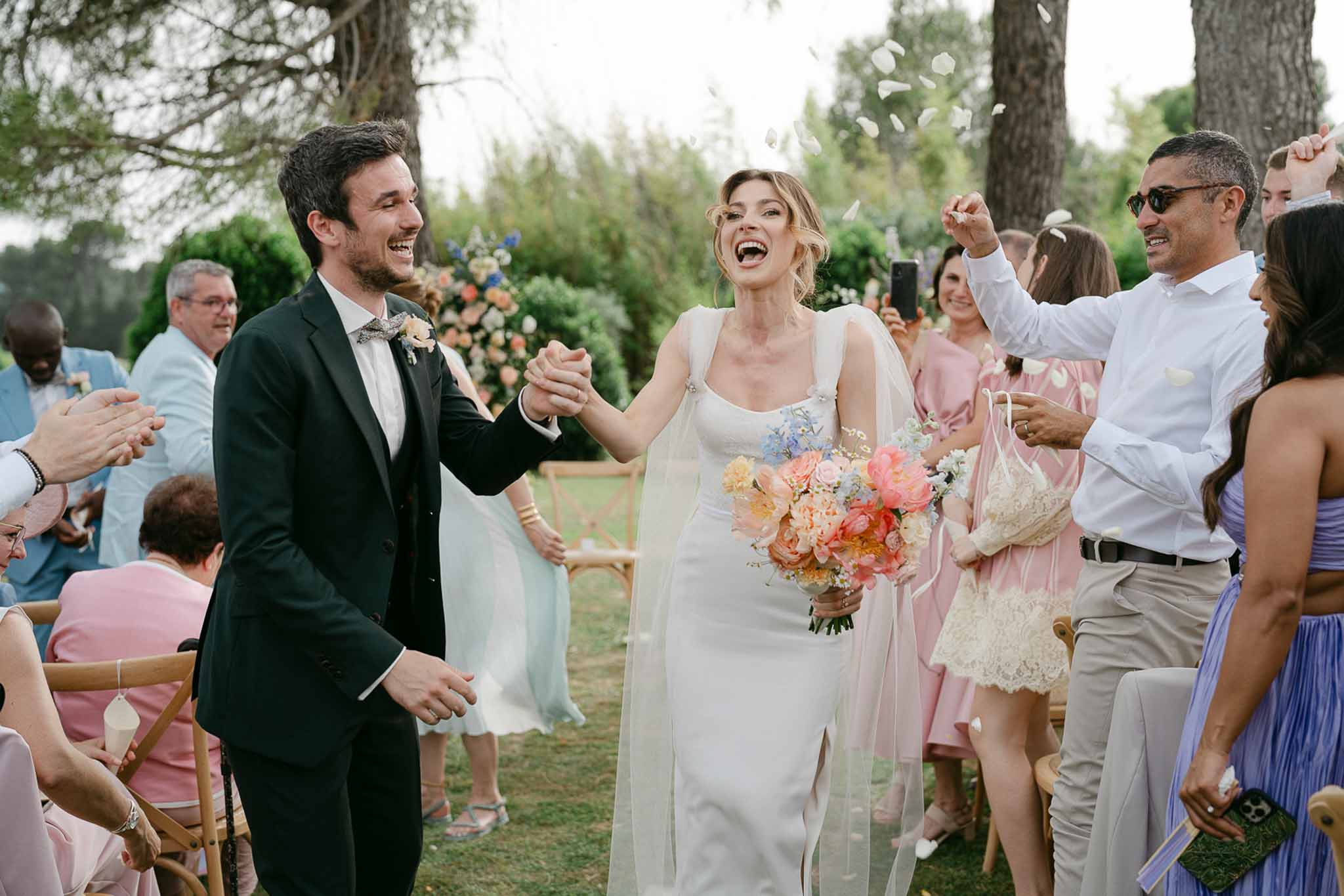 Bride and groom walking back down aisle as guests throw petals, pink bridesmaids and blue groomsmen visible on either side