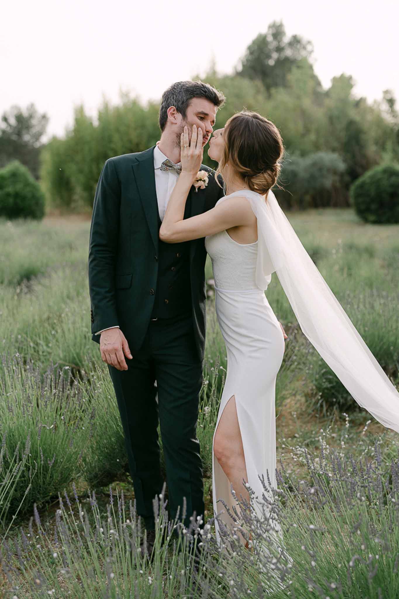 Bride kissing groom's cheek in lavender field wearing ivory column gown with flowing veil