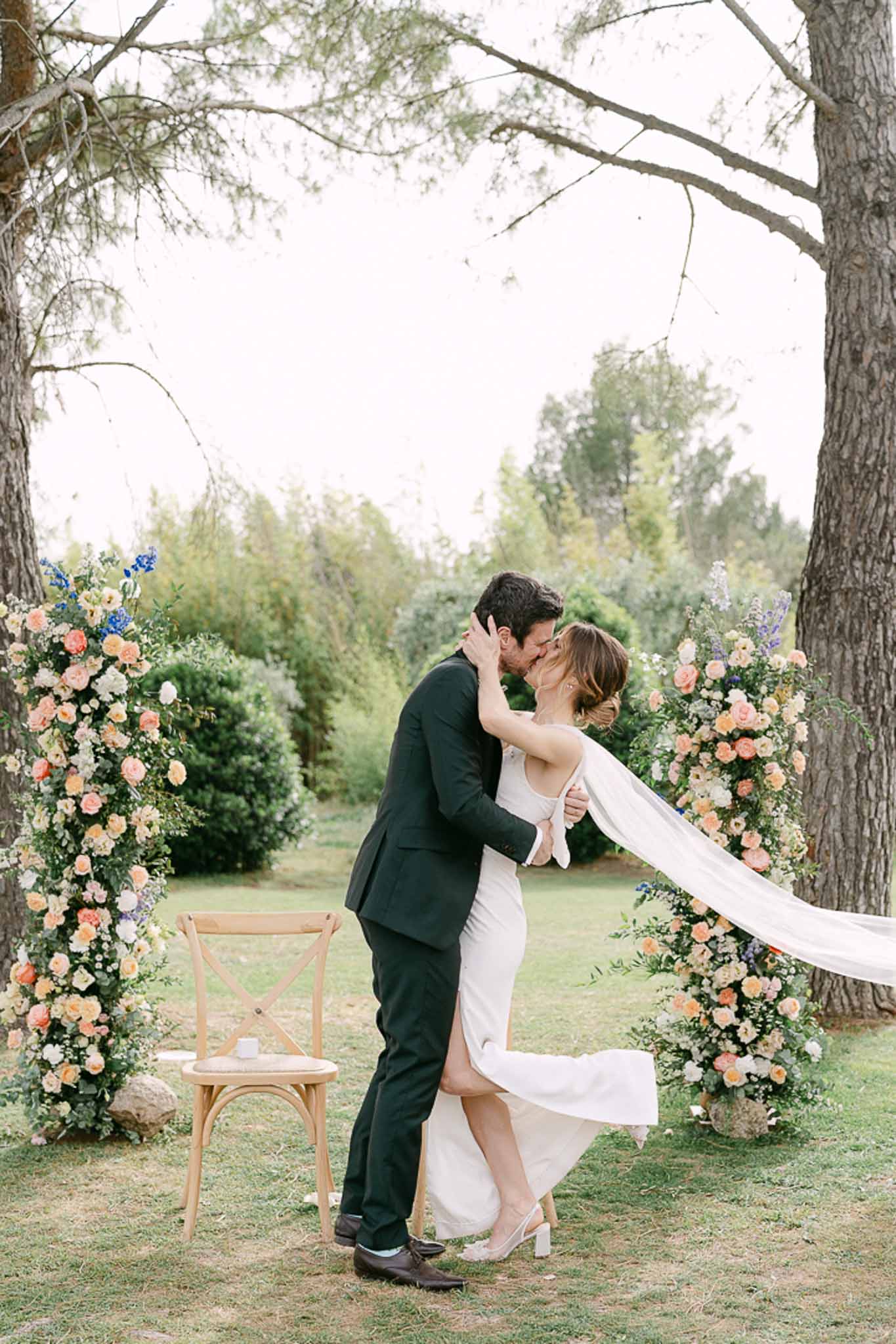 Bride and groom kiss beneath a floral arch of apricot roses and blue delphinium in an outdoor garden setting