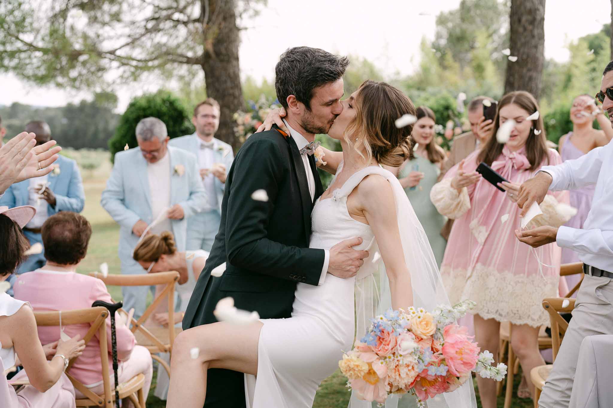 Bride and groom kissing at ceremony as guests applaud in outdoor garden with cypress trees, coral and peach bridal bouquet