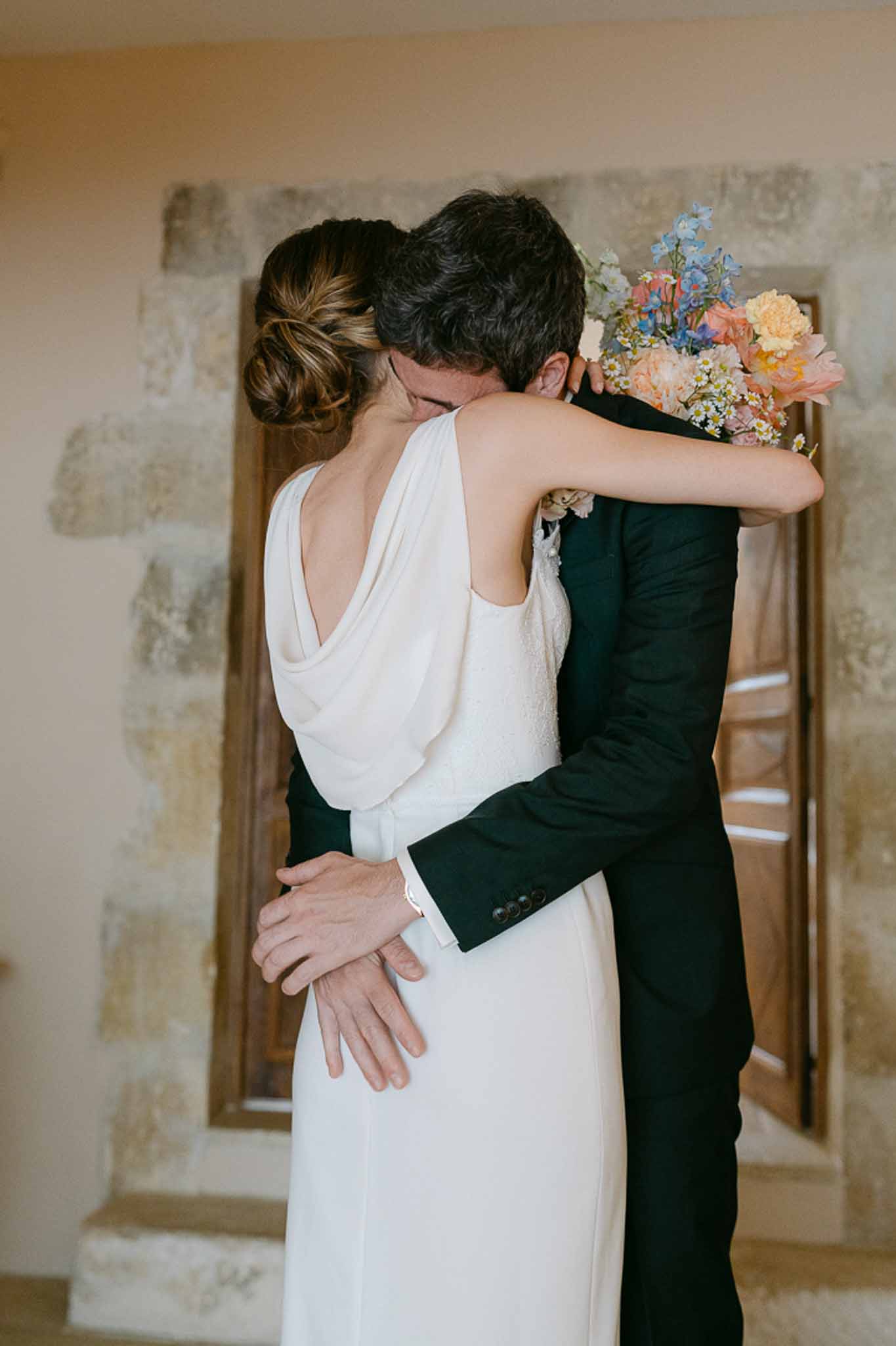 Bride and groom embrace indoors, bride holding a mixed bouquet of coral peonies and eucalyptus