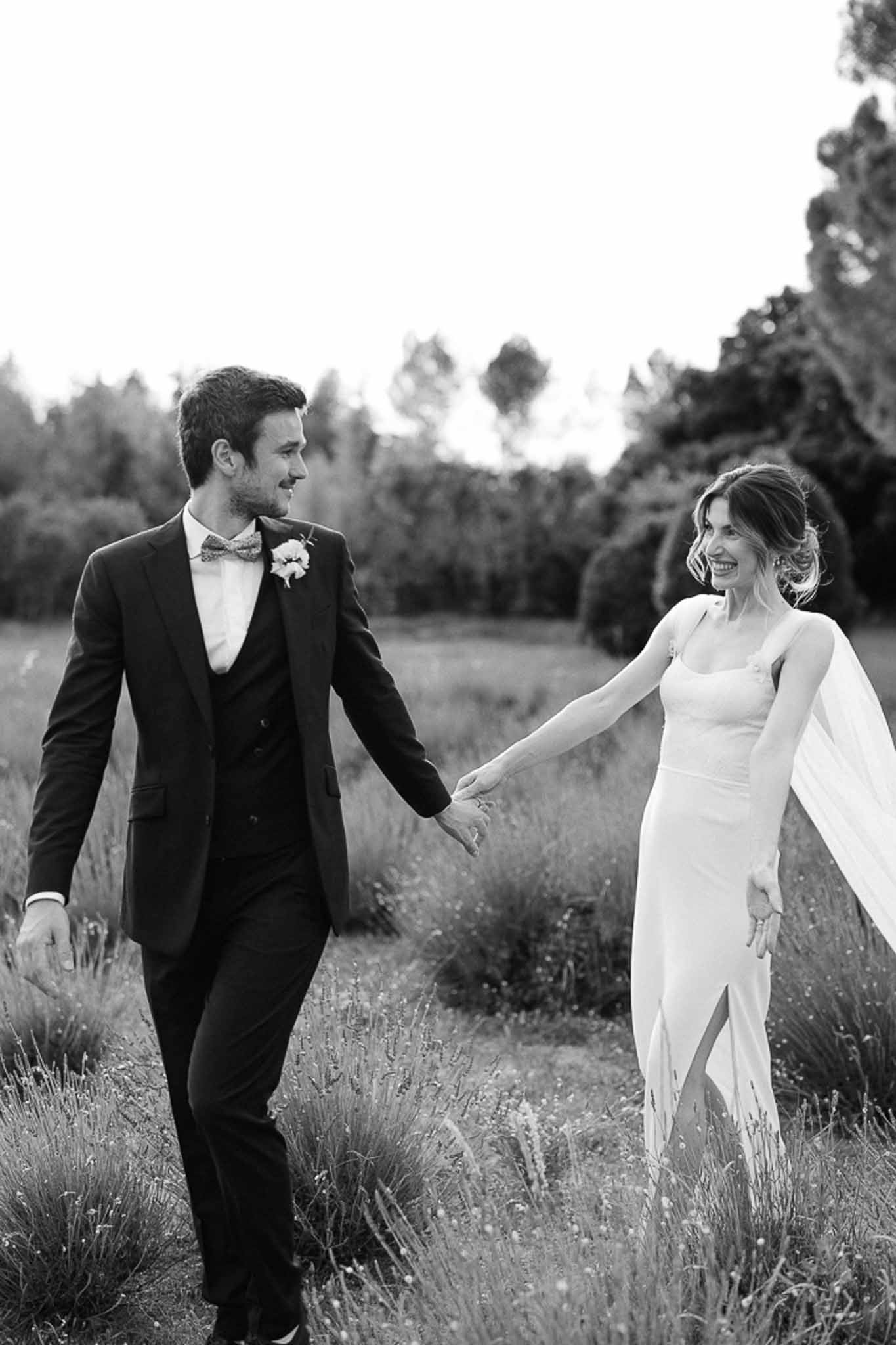 Black and white portrait of bride and groom walking hand-in-hand through open field smiling