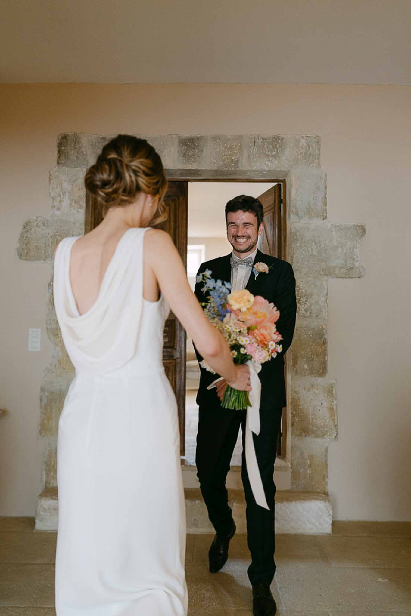 Groom approaching bride through a stone archway as she holds a coral dahlia and dusty miller bouquet