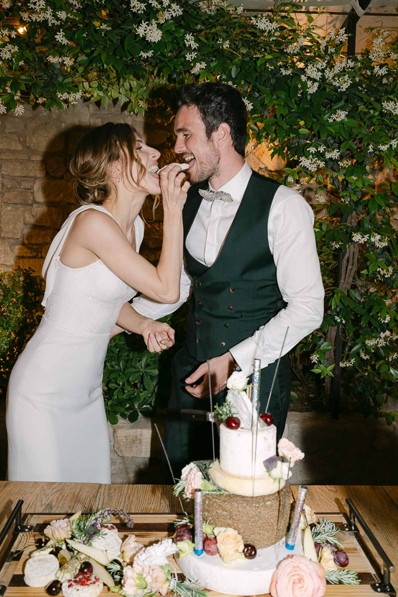 Couple feeding each other cake before jasmine-draped reception backdrop, two-tier white cake with blush roses