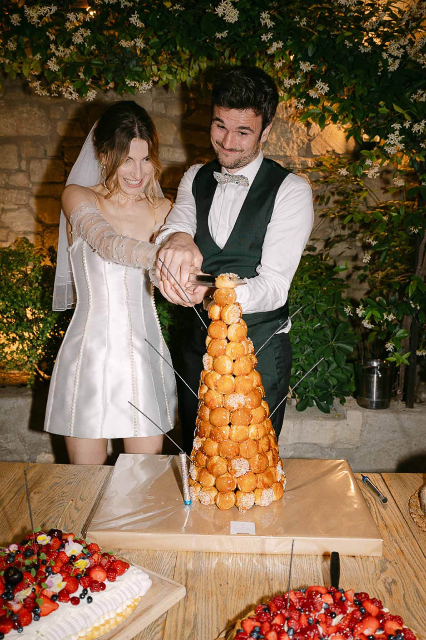 Bride and groom cut croquembouche at table before stone wall with climbing white florals at evening reception