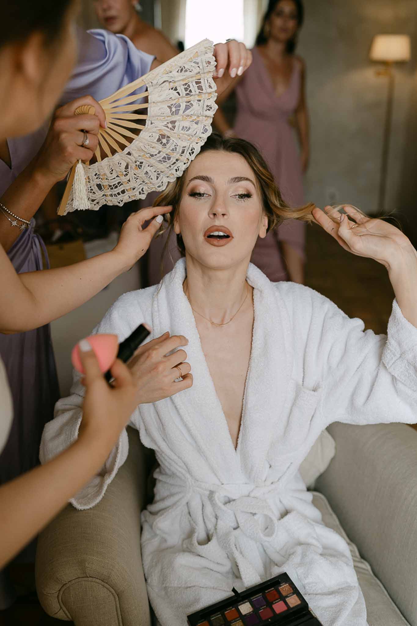 Bride in white robe seated while makeup is applied, bridesmaids in mauve and dusty rose robes visible behind her