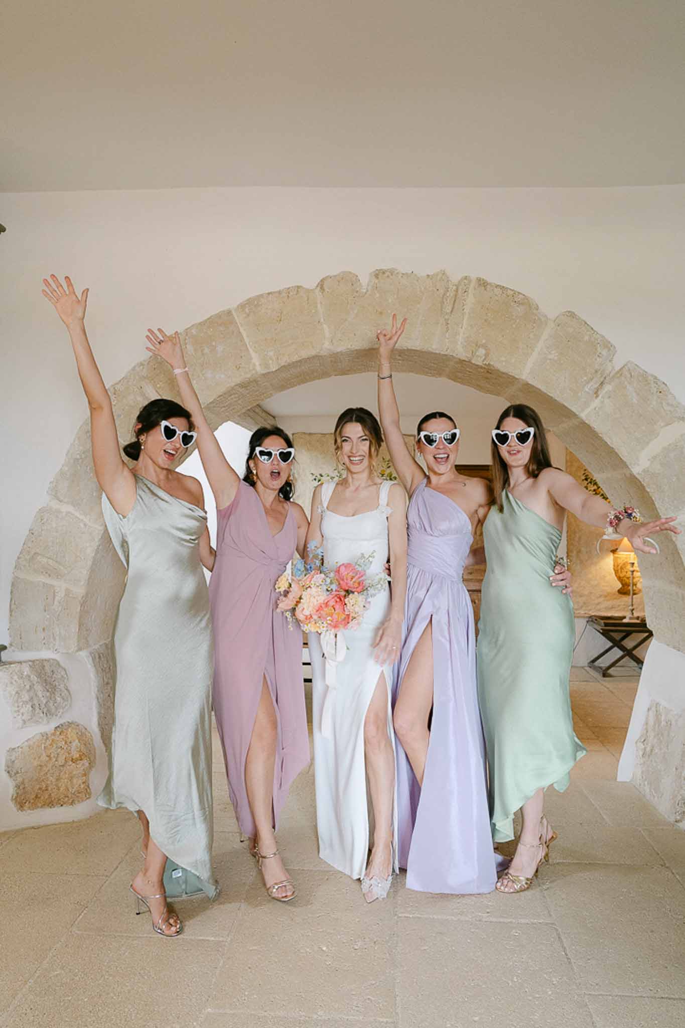 Bride and four bridesmaids in pastel dresses with heart sunglasses under stone archway