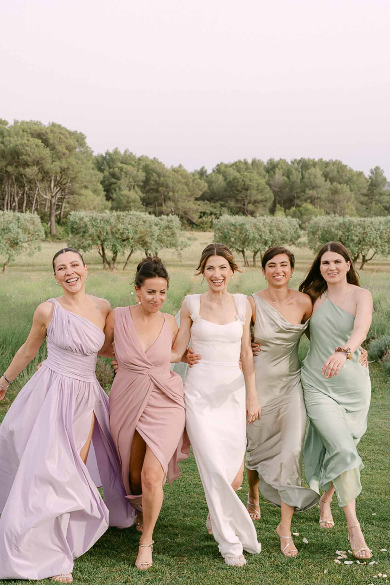 Bride in ivory cowl-neck gown with four bridesmaids in mismatched muted dresses on manicured lawn with olive trees