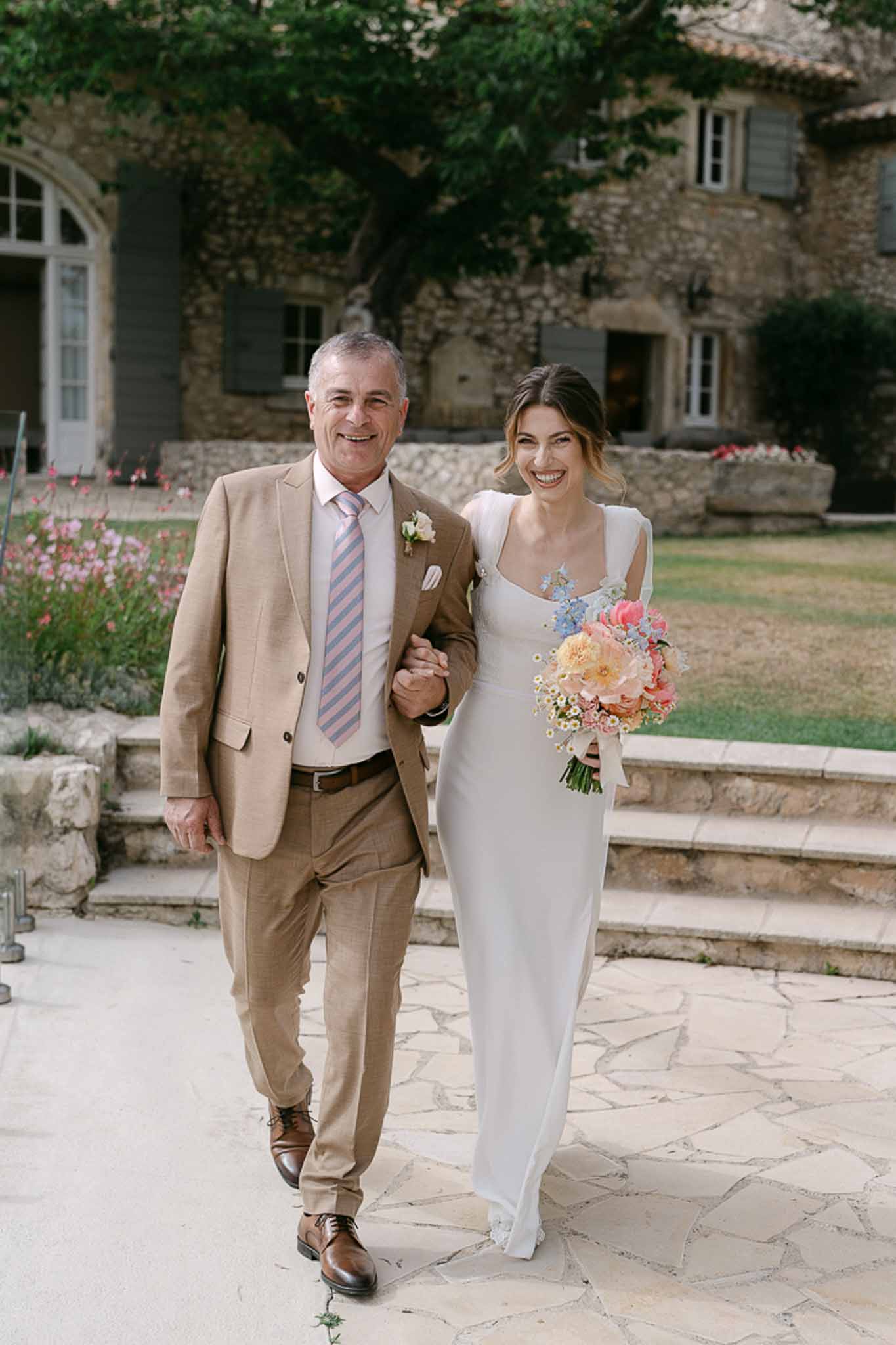 Bride in ivory dress with peach and blue bouquet walks arm-in-arm with father in beige suit across stone courtyard